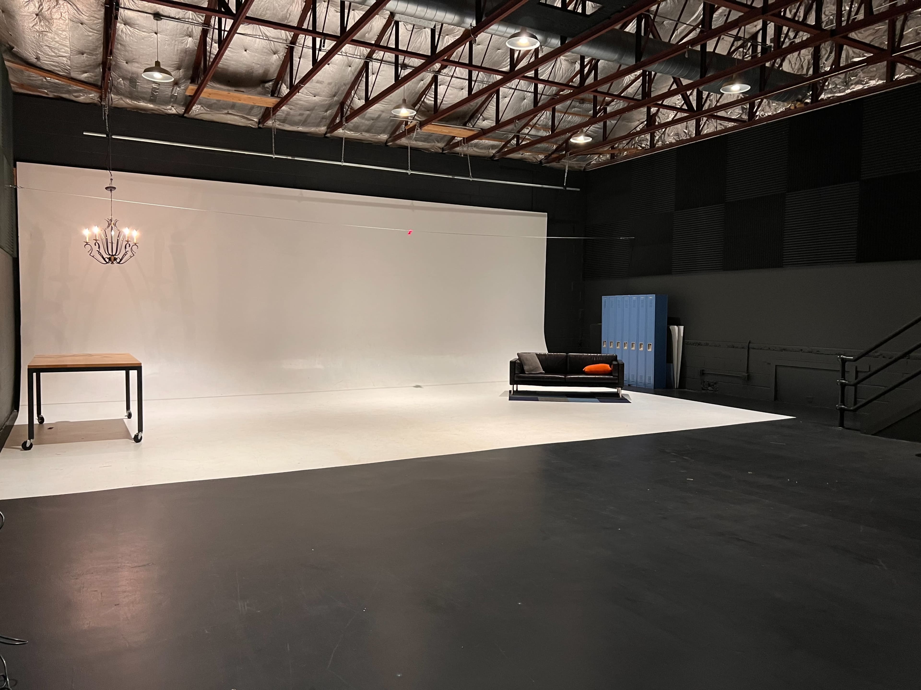 A spacious studio with a white backdrop, a black couch with an orange cushion, a wooden table on wheels, a chandelier, and blue lockers against a dark wall.