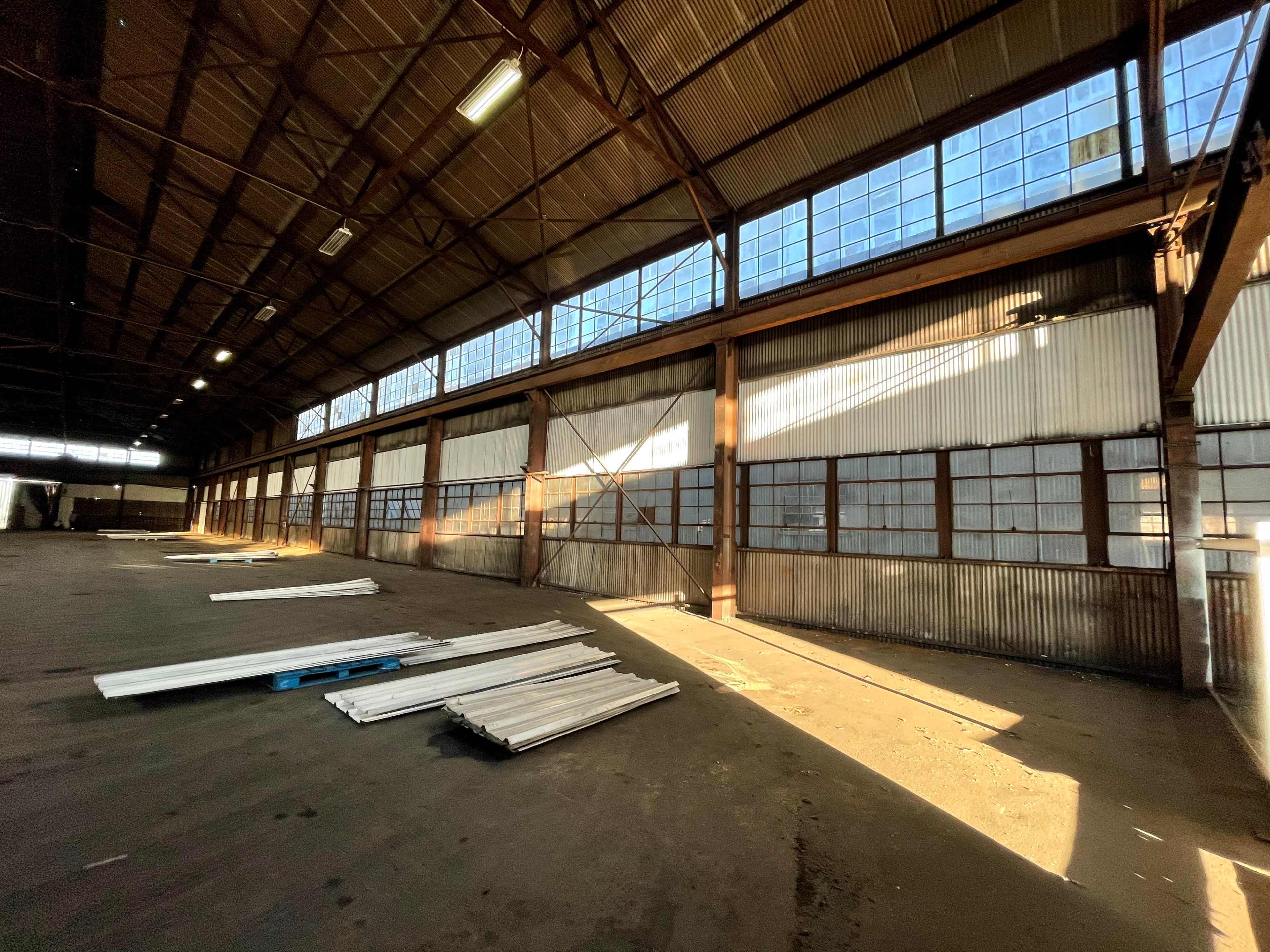 The interior of a large, empty warehouse with rows of windows along one wall and metal panels lining the opposite wall, while materials are scattered on the floor.