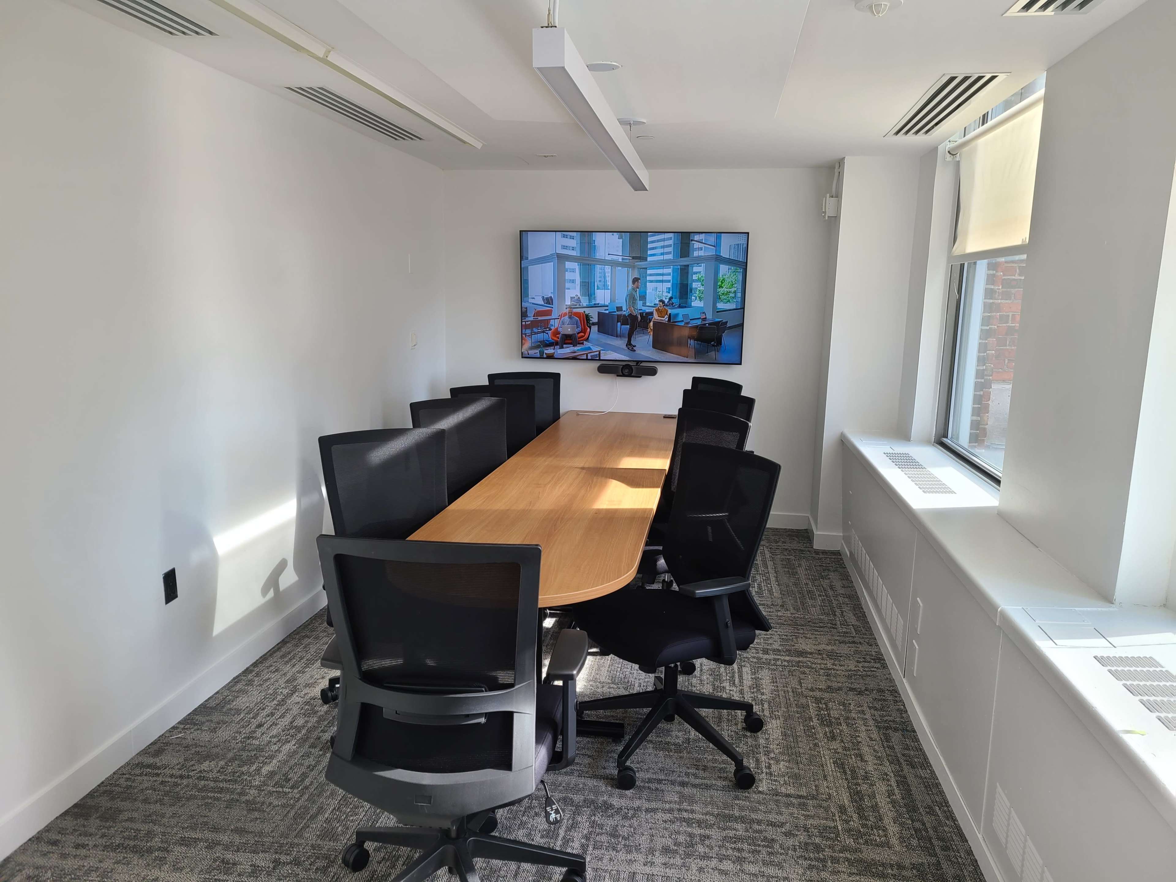 A modern conference room features a long wooden table surrounded by black ergonomic chairs, with a large screen mounted on the wall displaying a bright, airy interior.