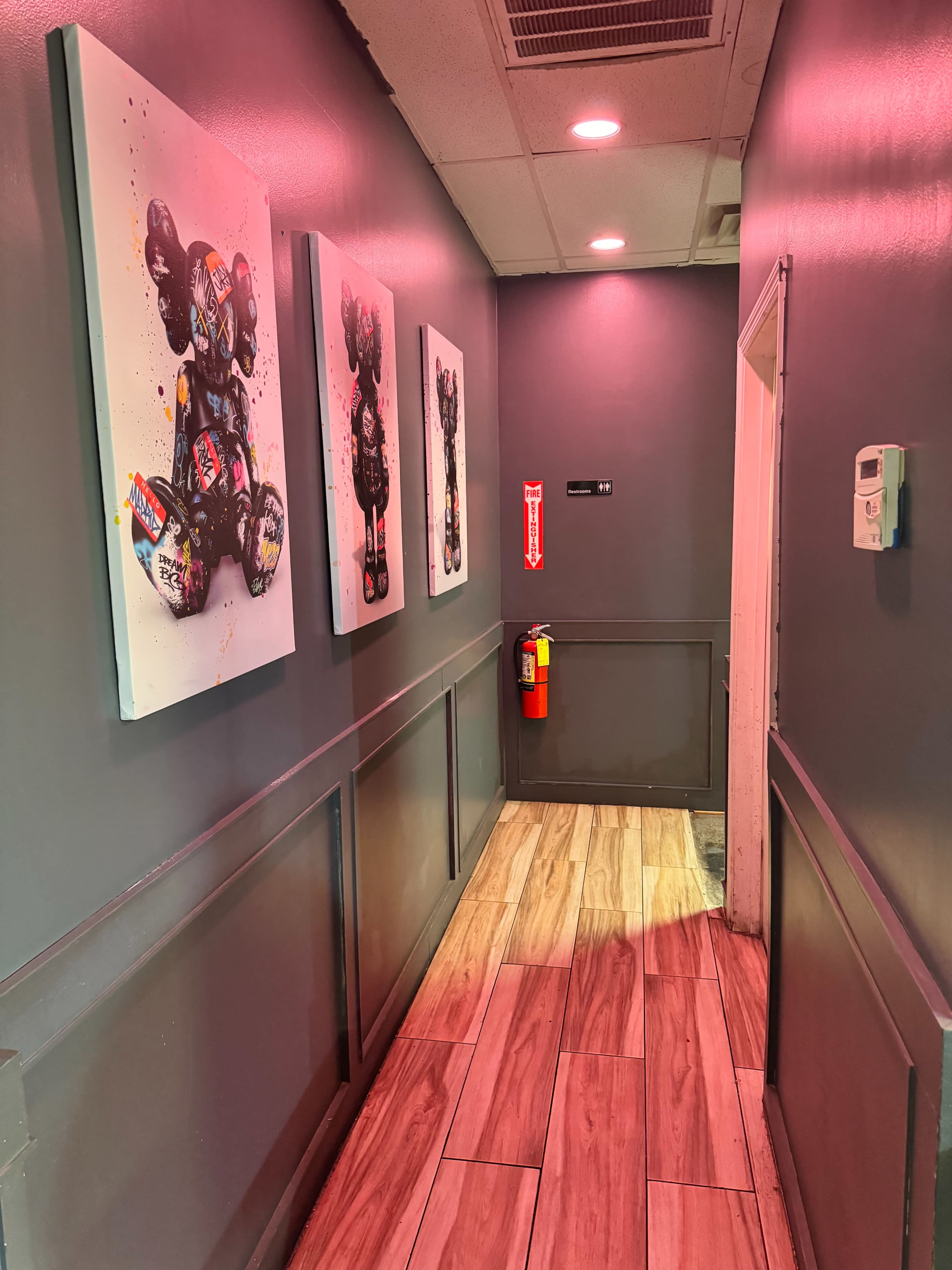 The narrow hallway features three colorful artworks of bear-like figures on the walls, with a fire extinguisher mounted next to a door at the end.