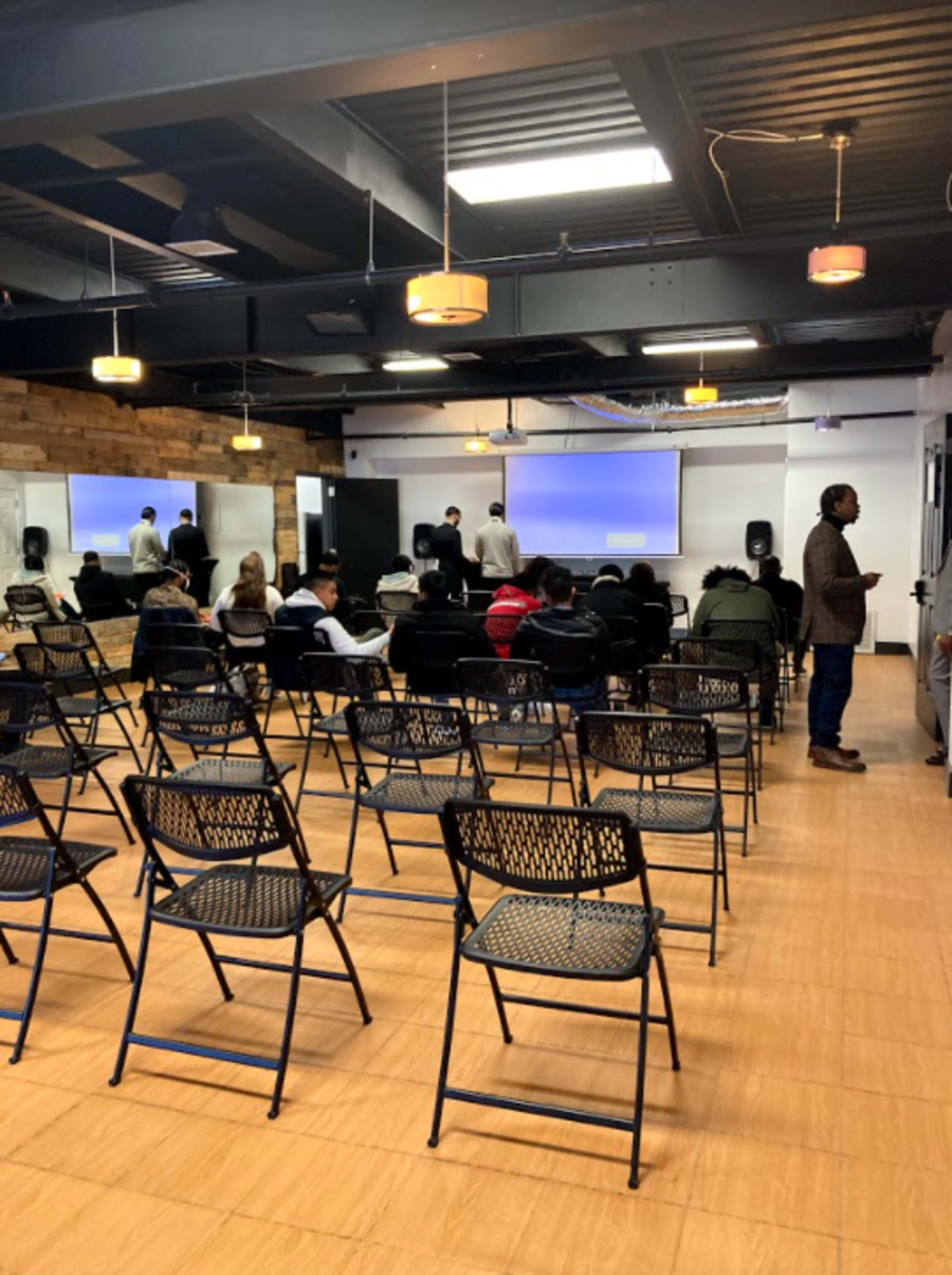 A group of people is seated in a classroom with empty chairs arranged in rows facing two screens at the front.