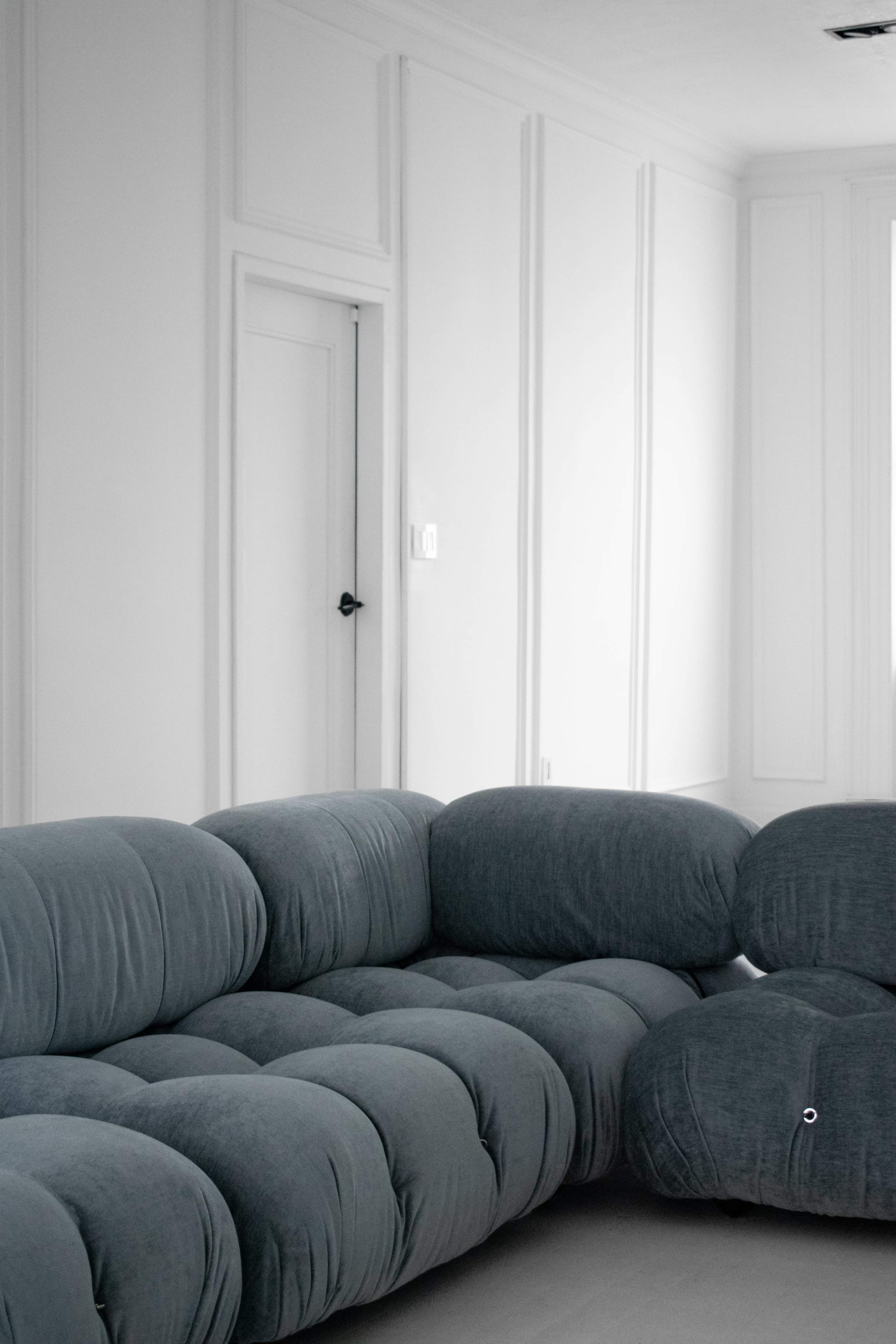 A gray upholstered sectional sofa is positioned in a minimalist room with white walls and a closed door.