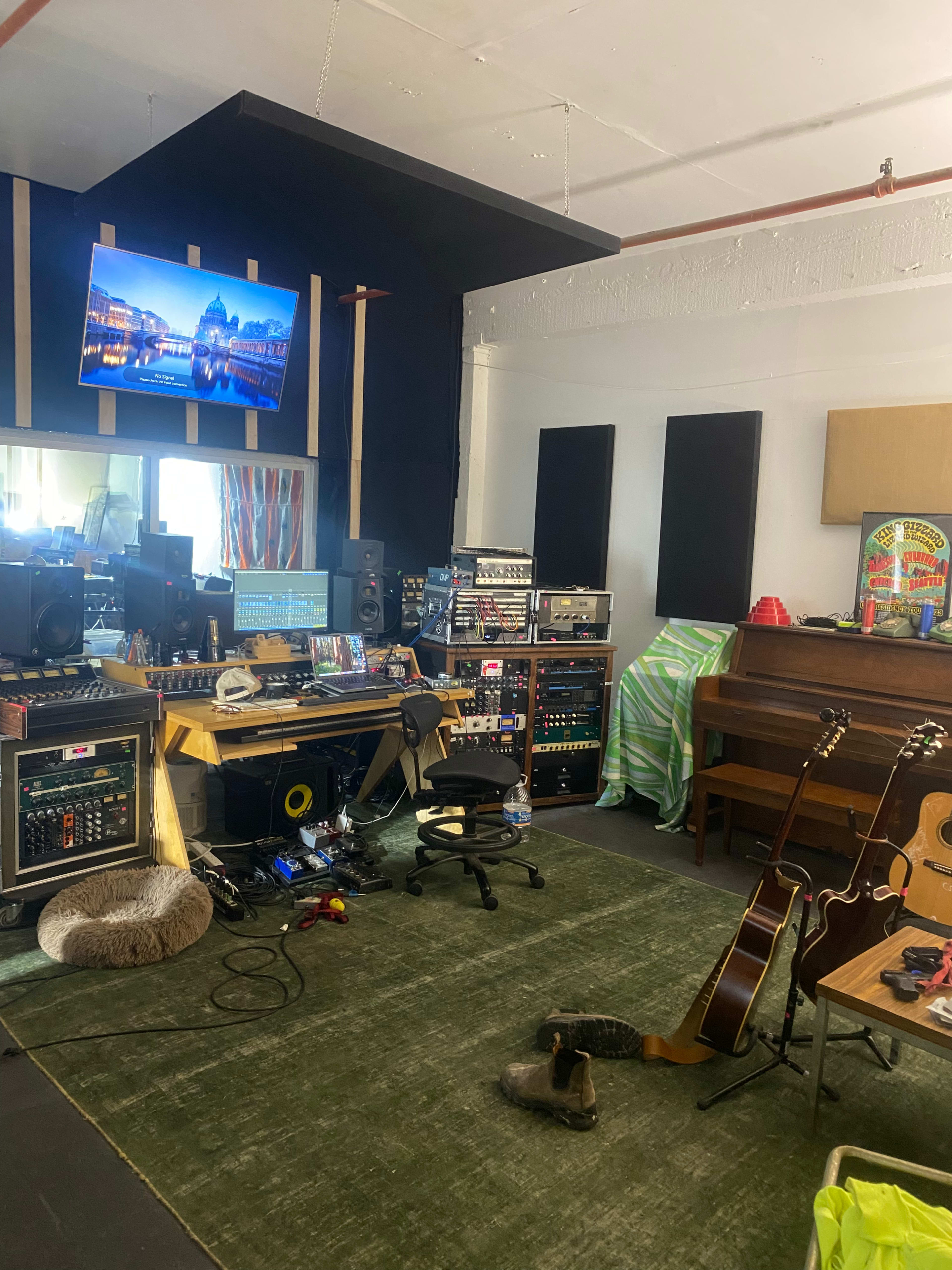 The image shows a music studio featuring a large desk with audio equipment, a television mounted on the wall, various instruments including guitars, and a carpeted floor.