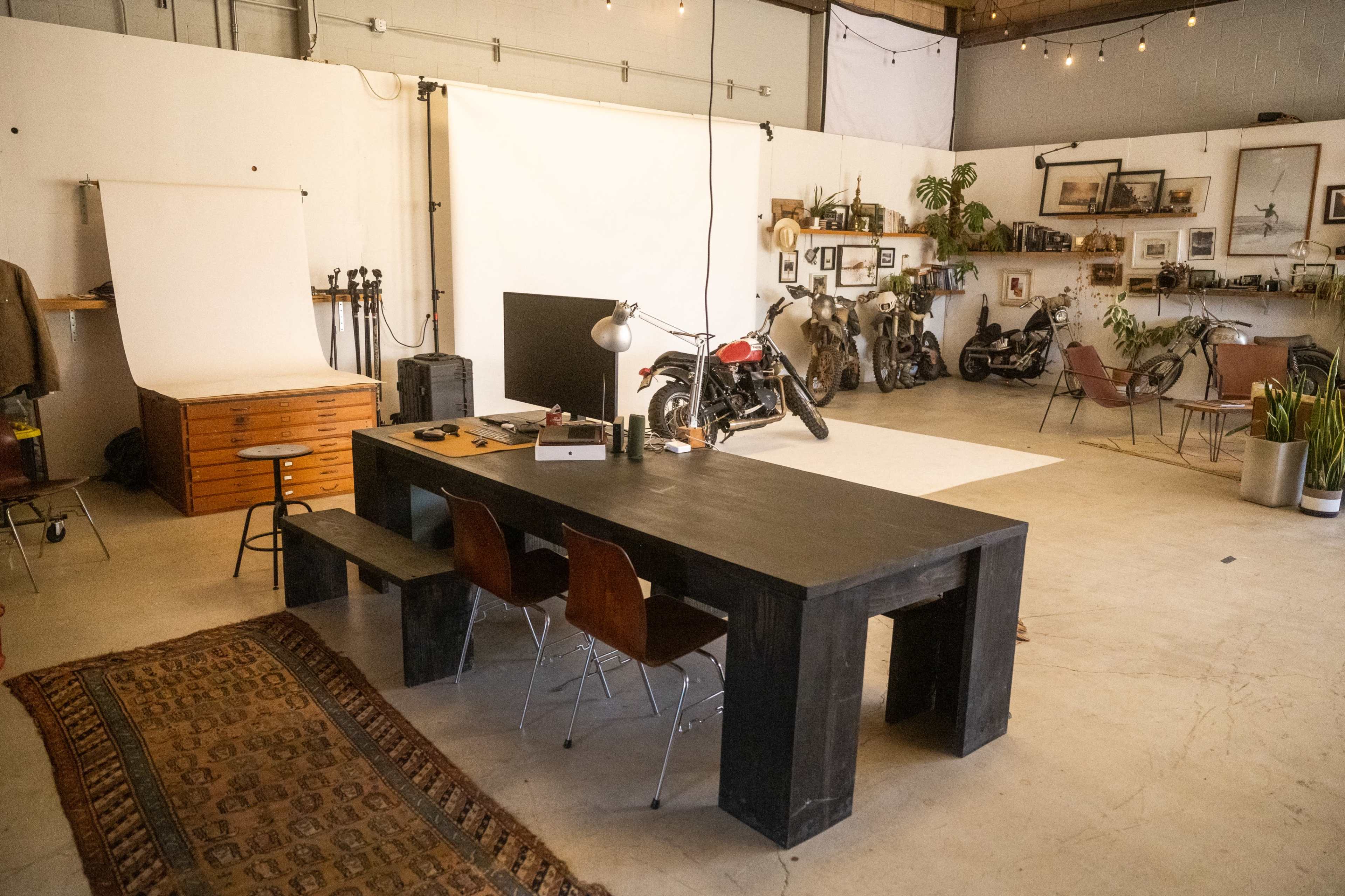 A spacious studio with a large black table, wooden chairs, a photography backdrop, vintage motorcycles, and various framed pictures on the walls.