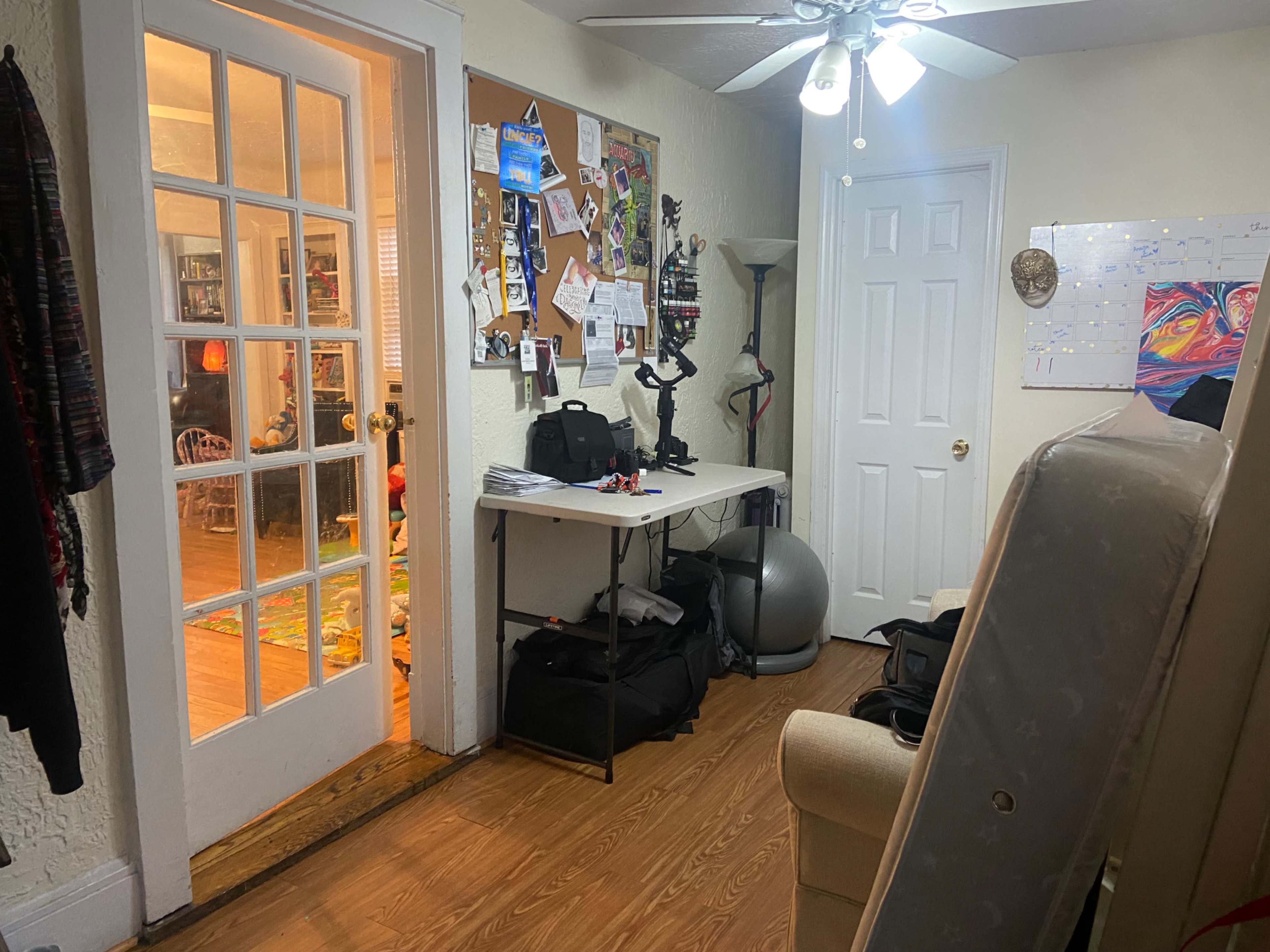 A hallway leads to a room with a bulletin board and various furniture items, including a table and exercise equipment.