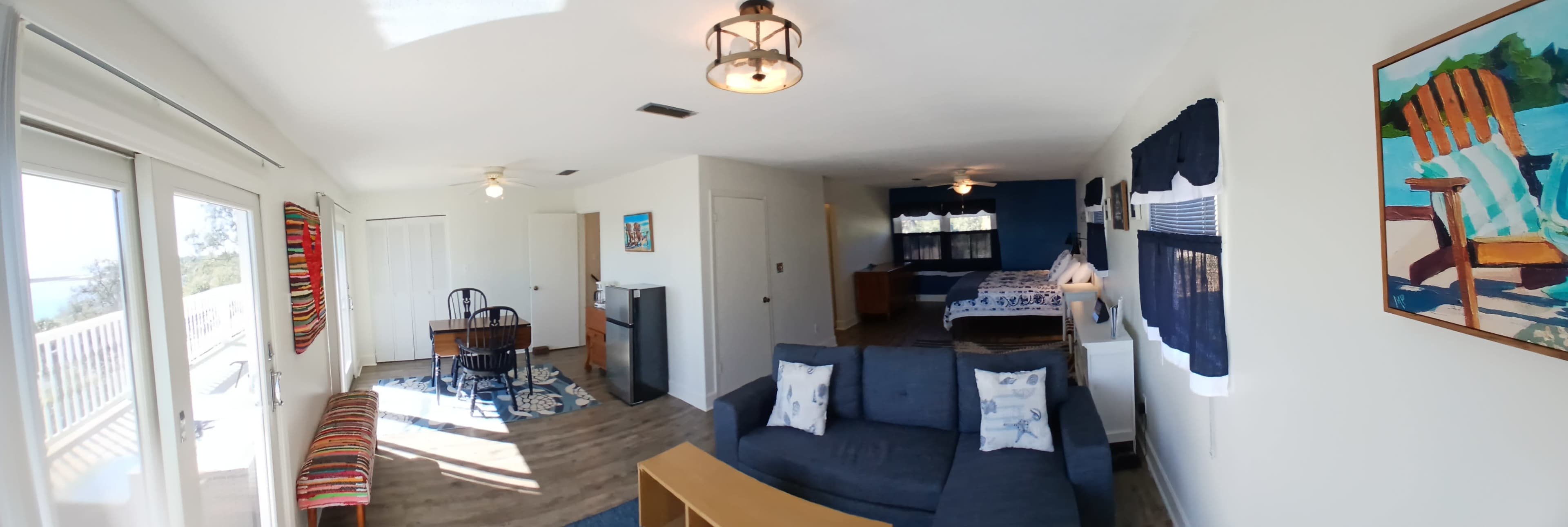 The image shows a bright, open-plan living space featuring a sitting area with a couch, a dining table, and a small kitchenette, leading to a bedroom area with a bed in the background.