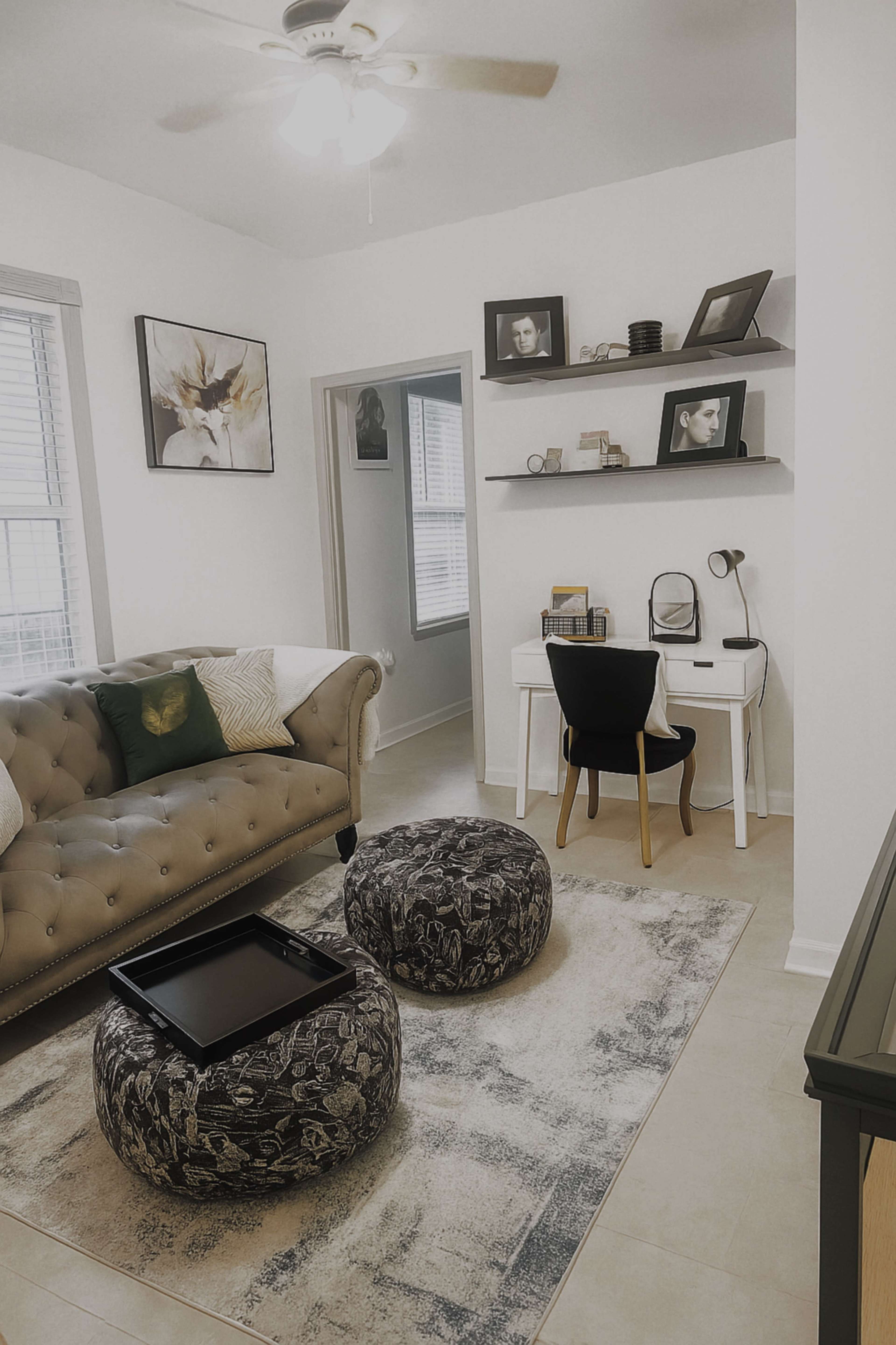 The image shows a well-furnished living space featuring a tufted couch, two patterned poufs, a desk with a chair, and wall-mounted shelves with framed pictures.