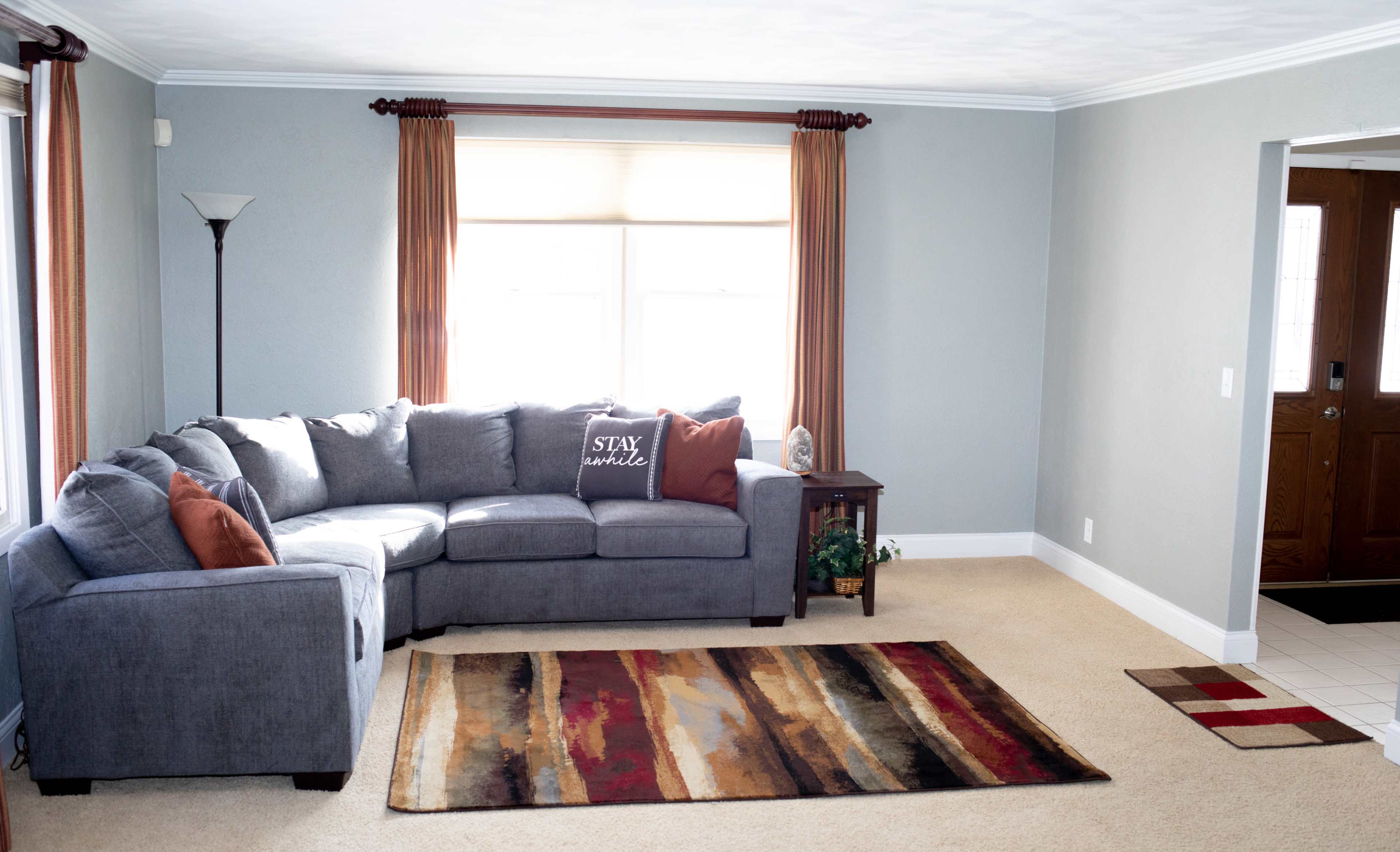 A gray sectional sofa with decorative pillows sits against a wall in a well-lit living room, accompanied by a multi-colored area rug and a wooden side table.