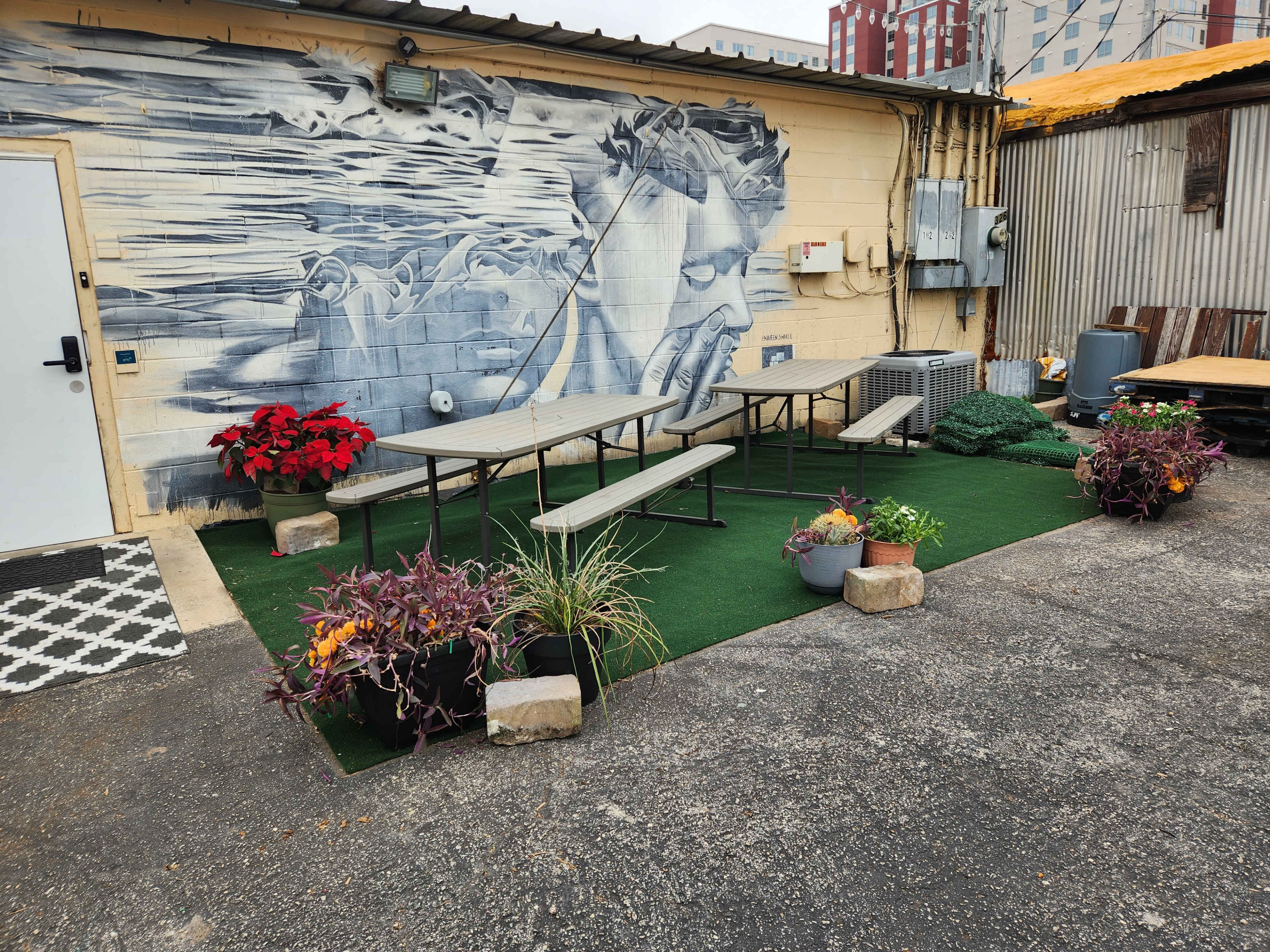 A small outdoor seating area with wooden tables and benches surrounded by potted plants and a mural on the wall.