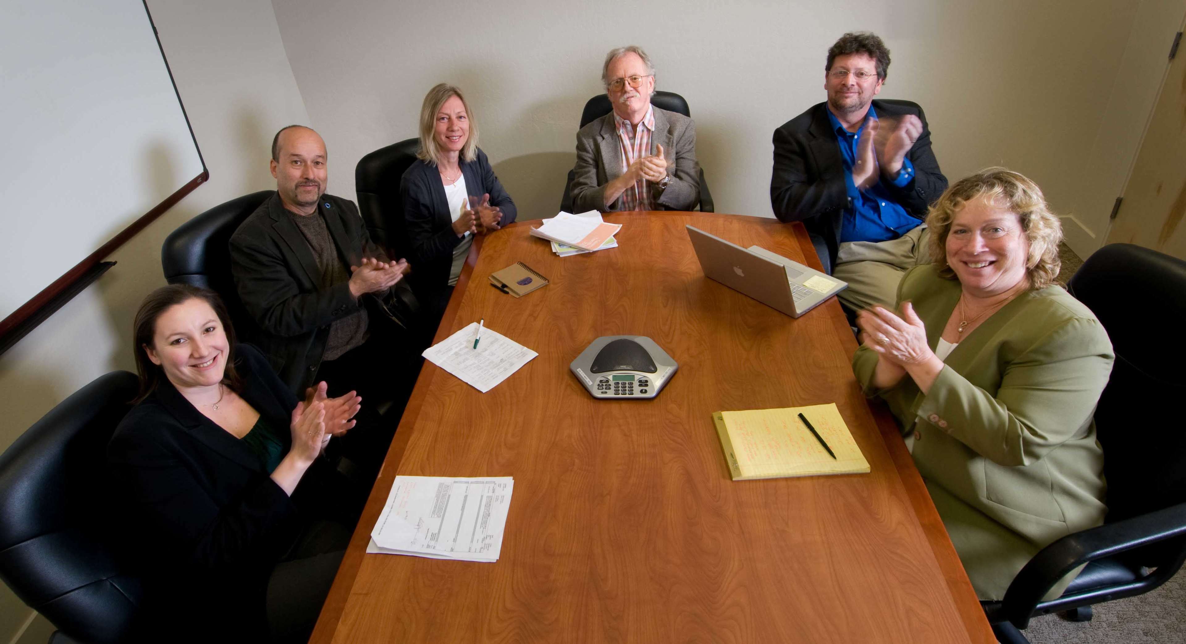 A group of six people is seated around a conference table, clapping their hands.