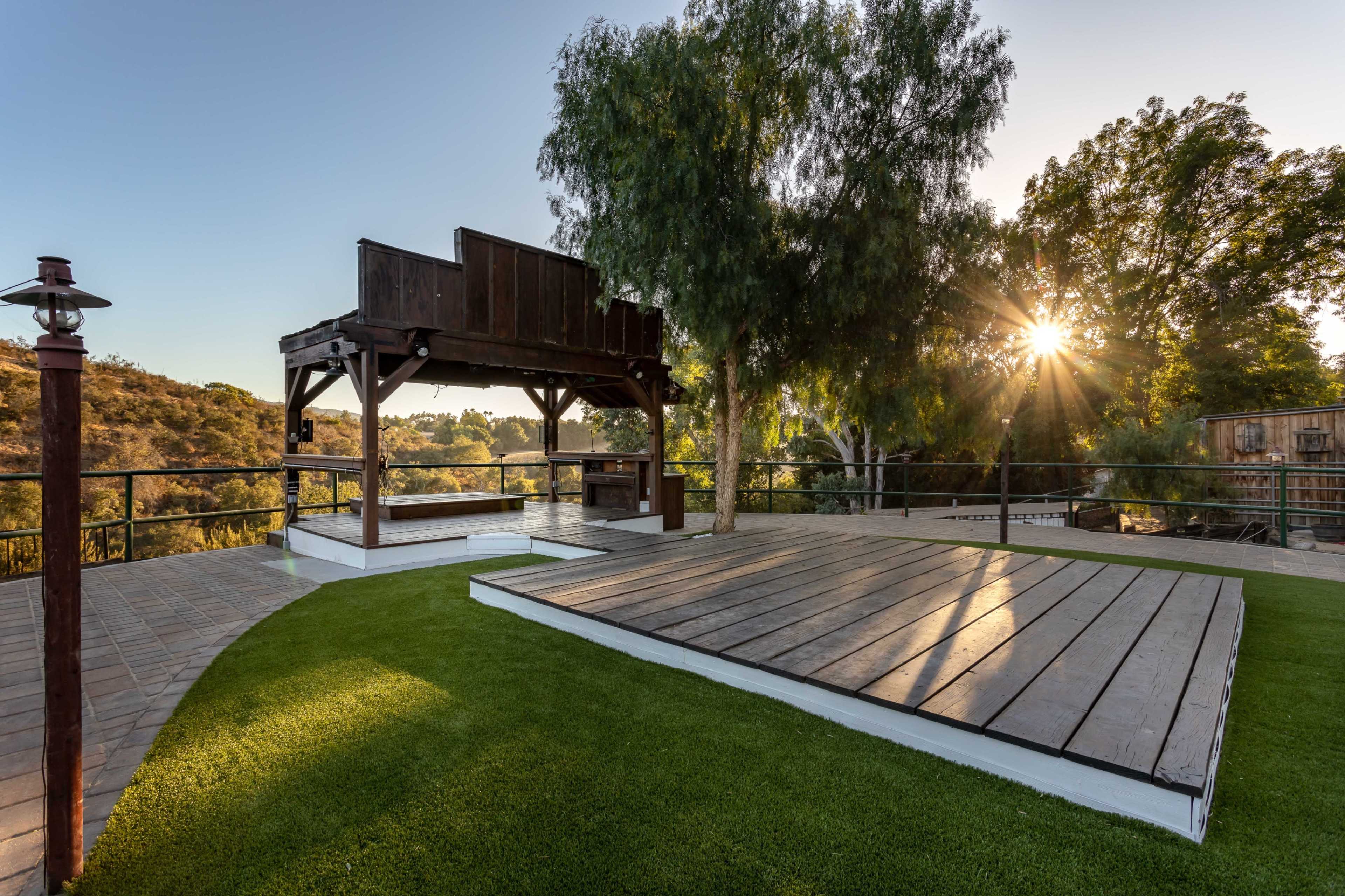 An outdoor space with a wooden pavilion, a flat deck area, and a view of trees and hills in the background, illuminated by sunlight.