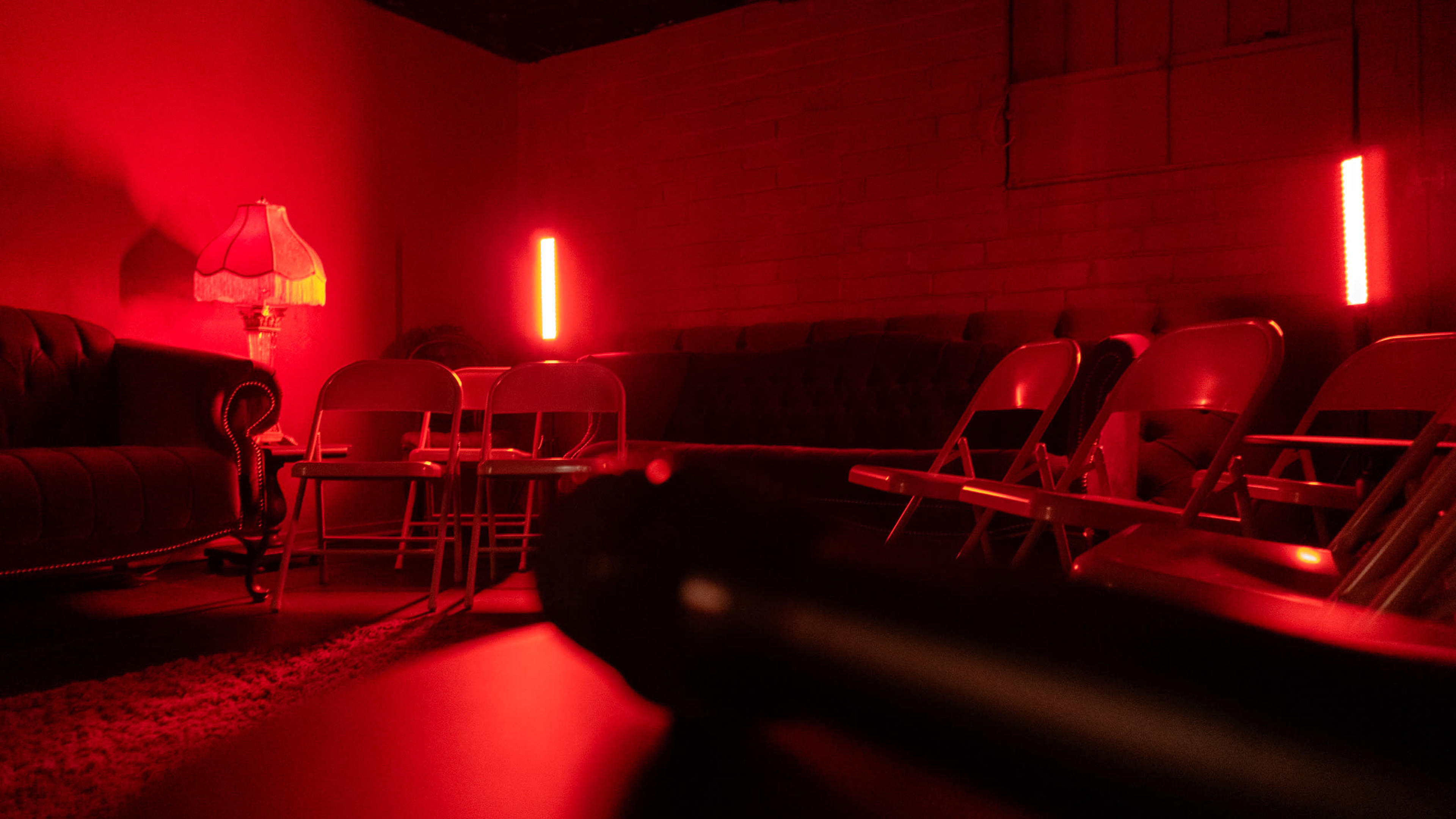 The image shows a dimly lit room with red lighting, featuring a couch, folding chairs, and a lamp.