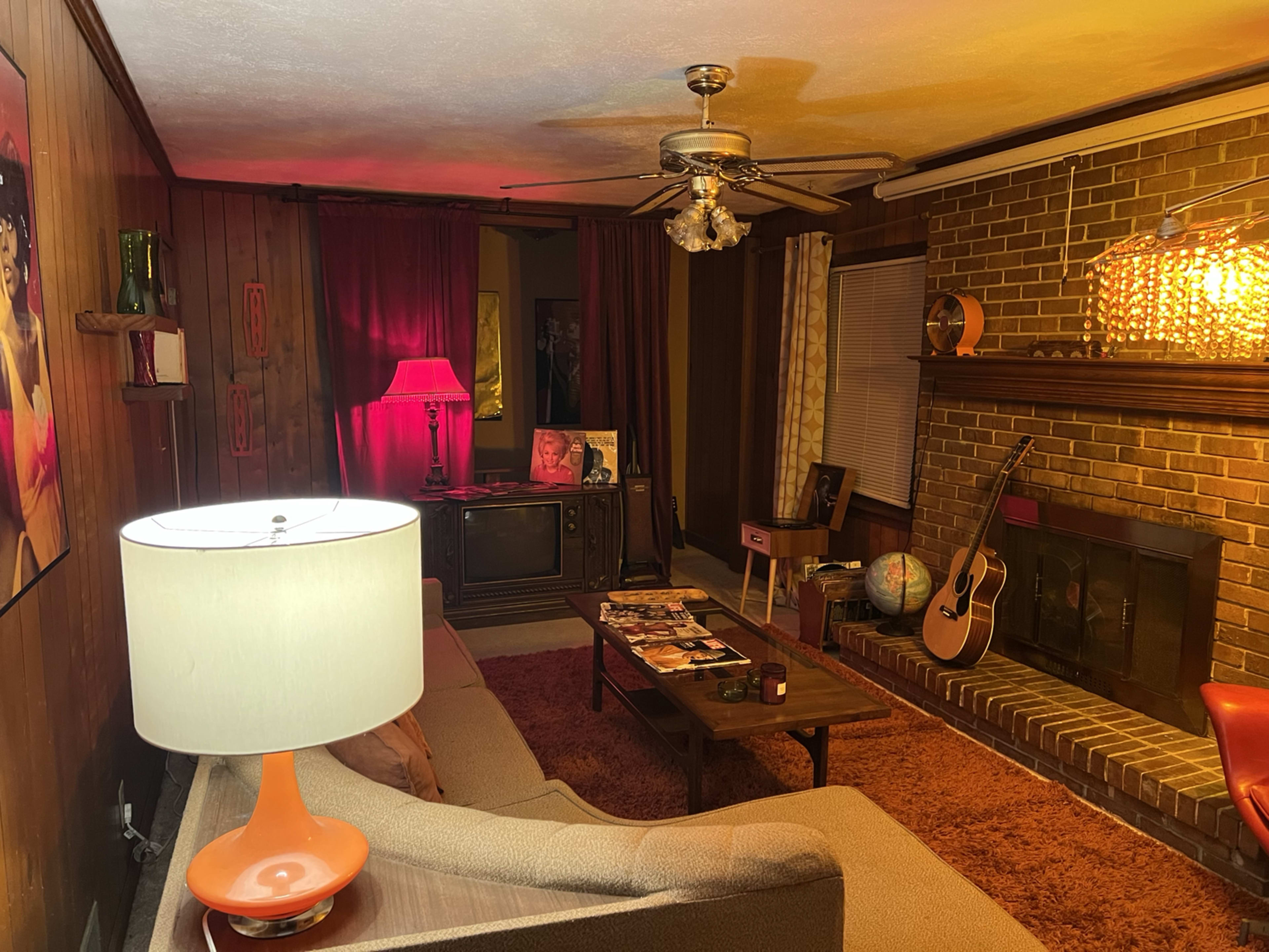 The living room features a vintage decor with a sofa, a lamp, a coffee table displaying magazines, and a fireplace surrounded by brick walls.
