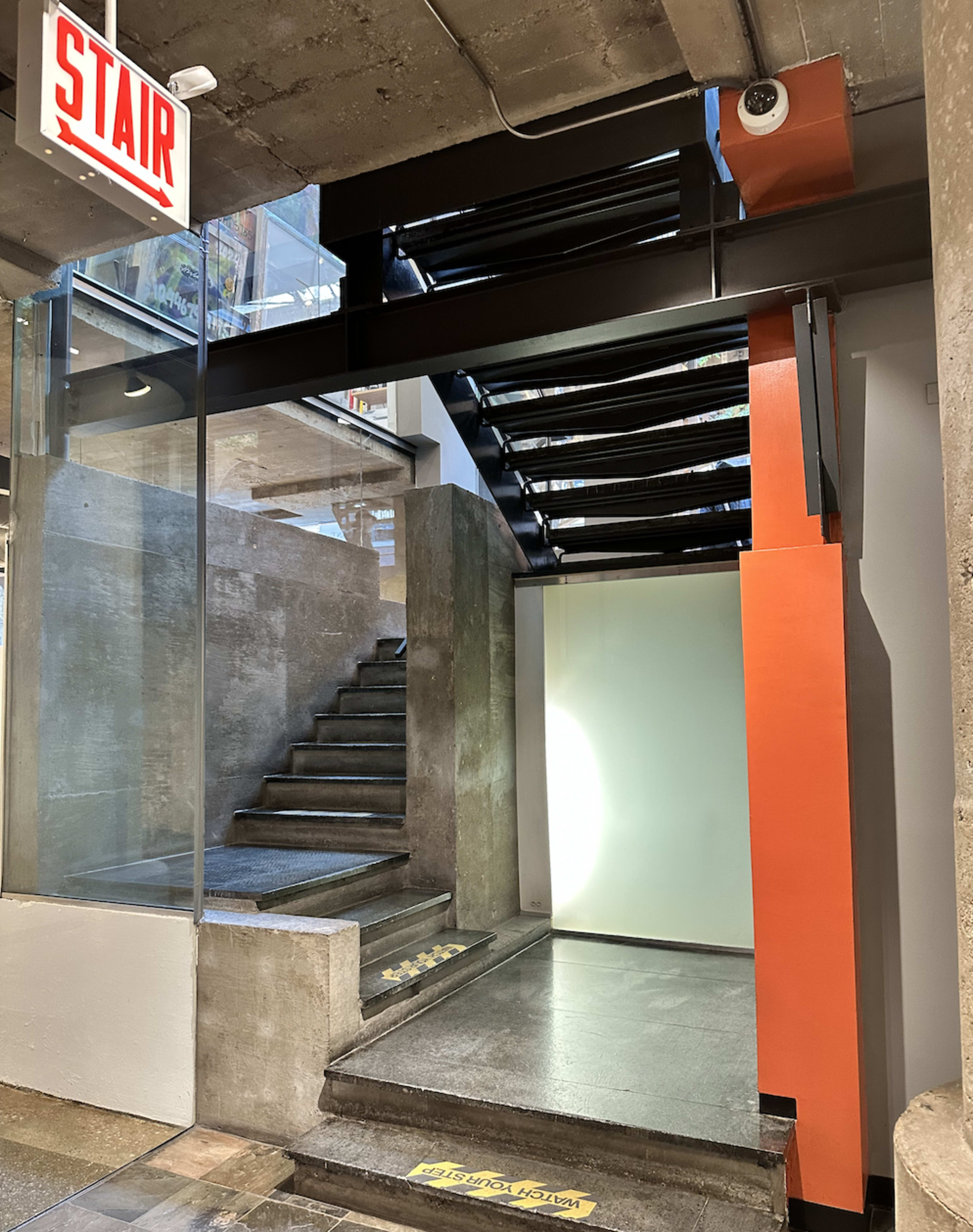 The image shows a modern interior featuring a staircase with a glass railing and an adjacent wall painted in orange, with a "STAIR" sign above.