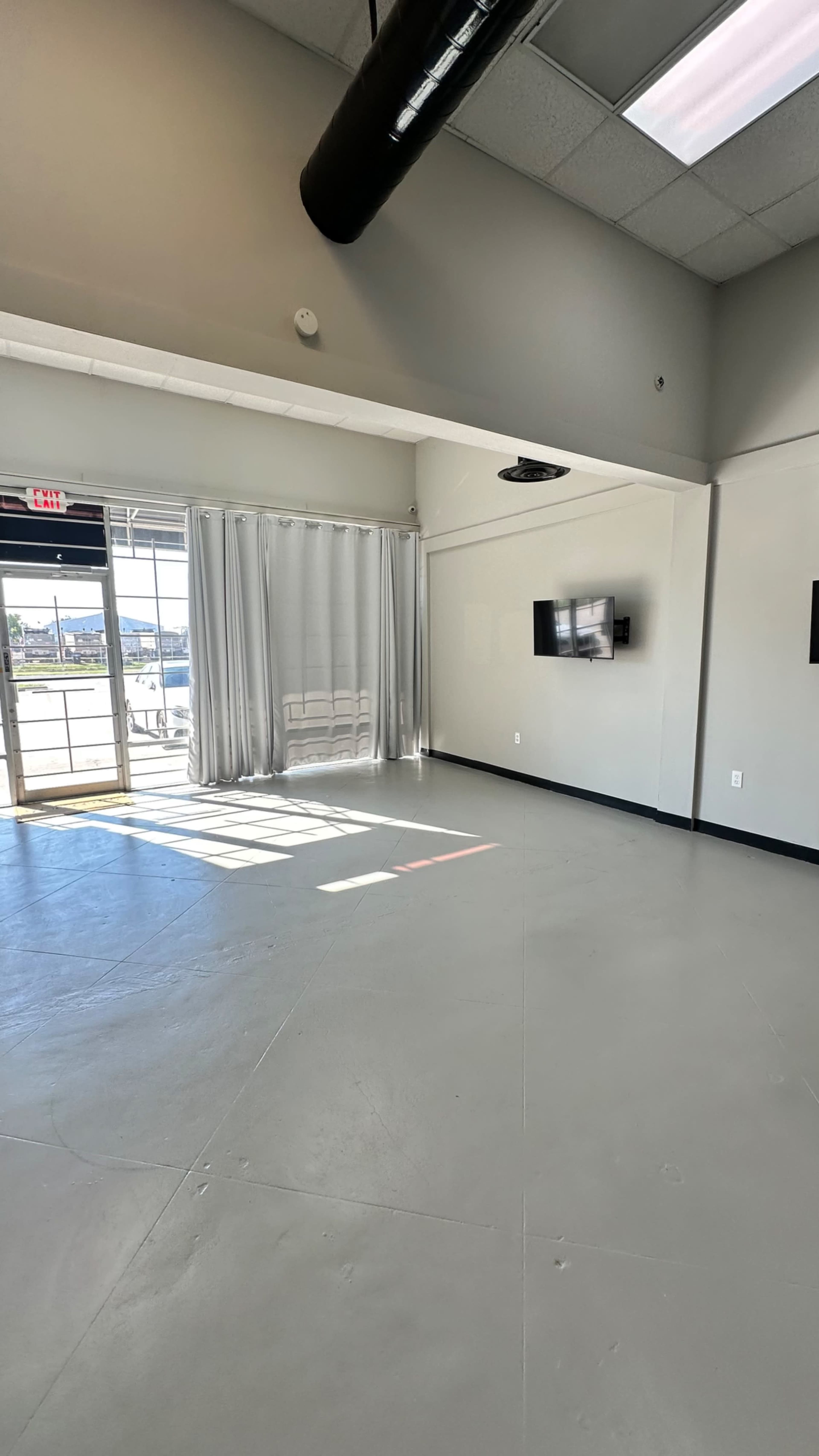 The image shows a spacious, empty room with gray flooring, large windows letting in natural light, and two wall-mounted televisions.
