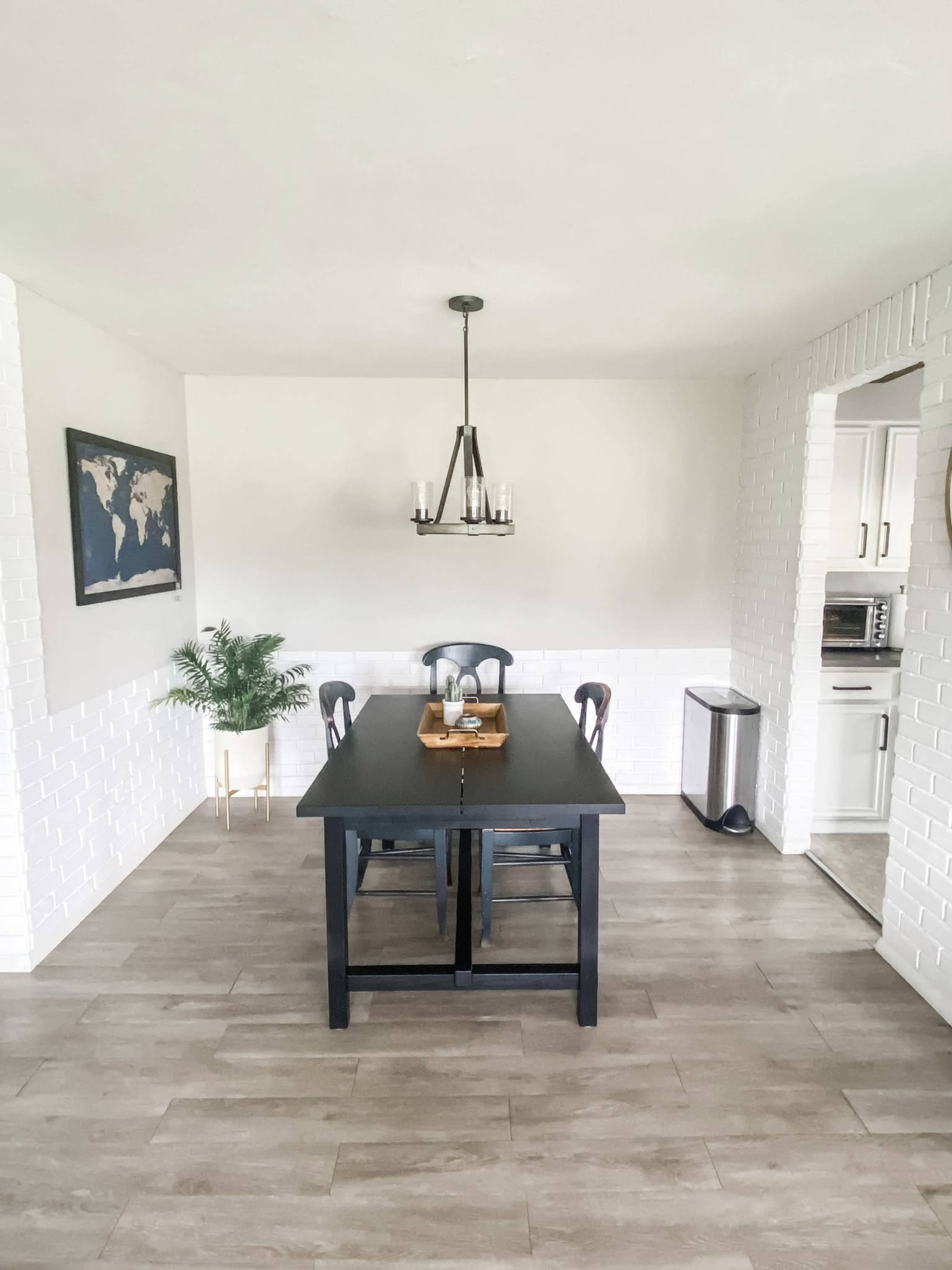 A dining area features a black rectangular table surrounded by four chairs, with a chandelier above and a brick wall on one side.