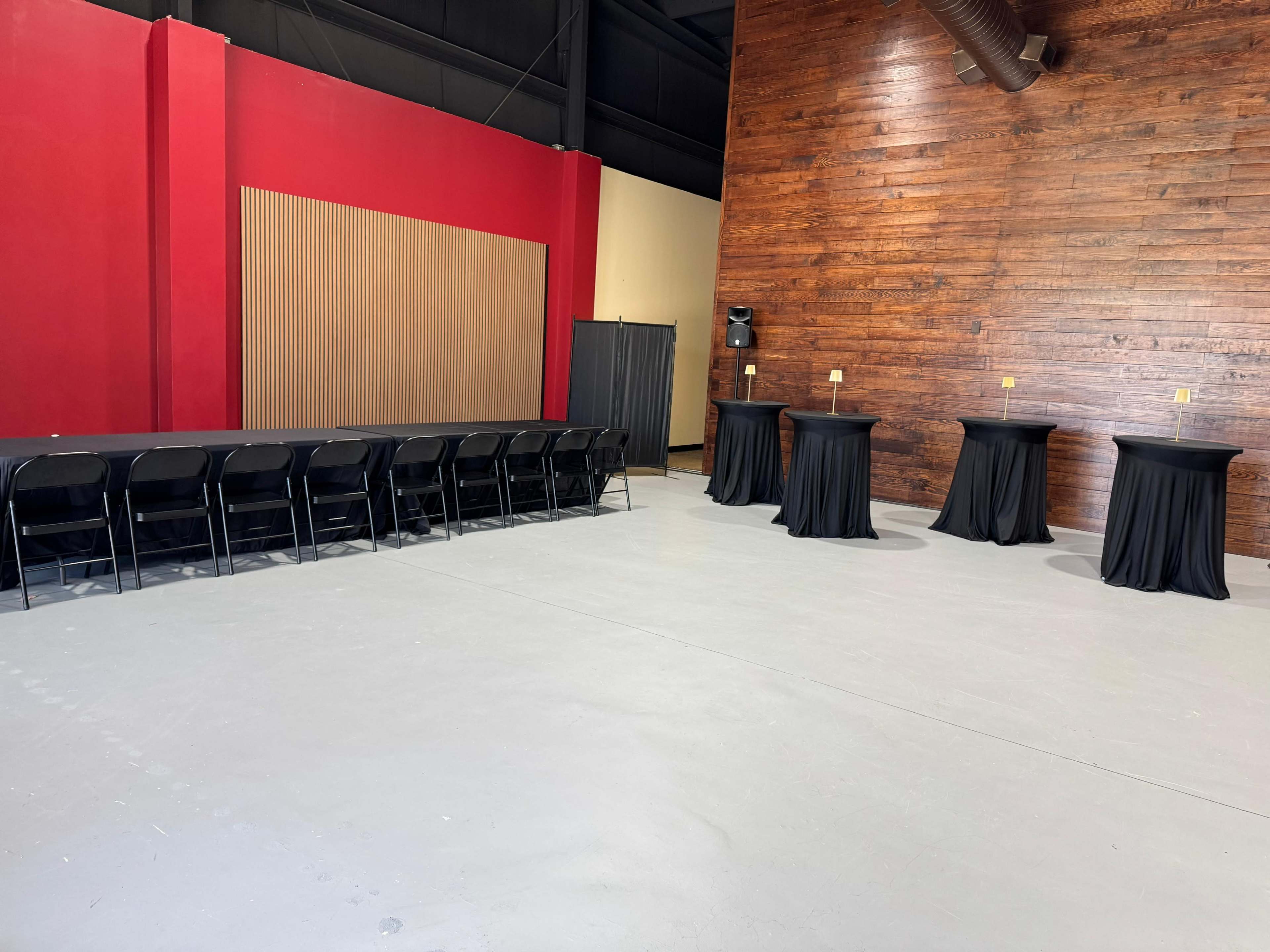 The image shows an empty indoor space with a long table lined with chairs on one side and several round tables with black tablecloths on the other side against a wooden wall.