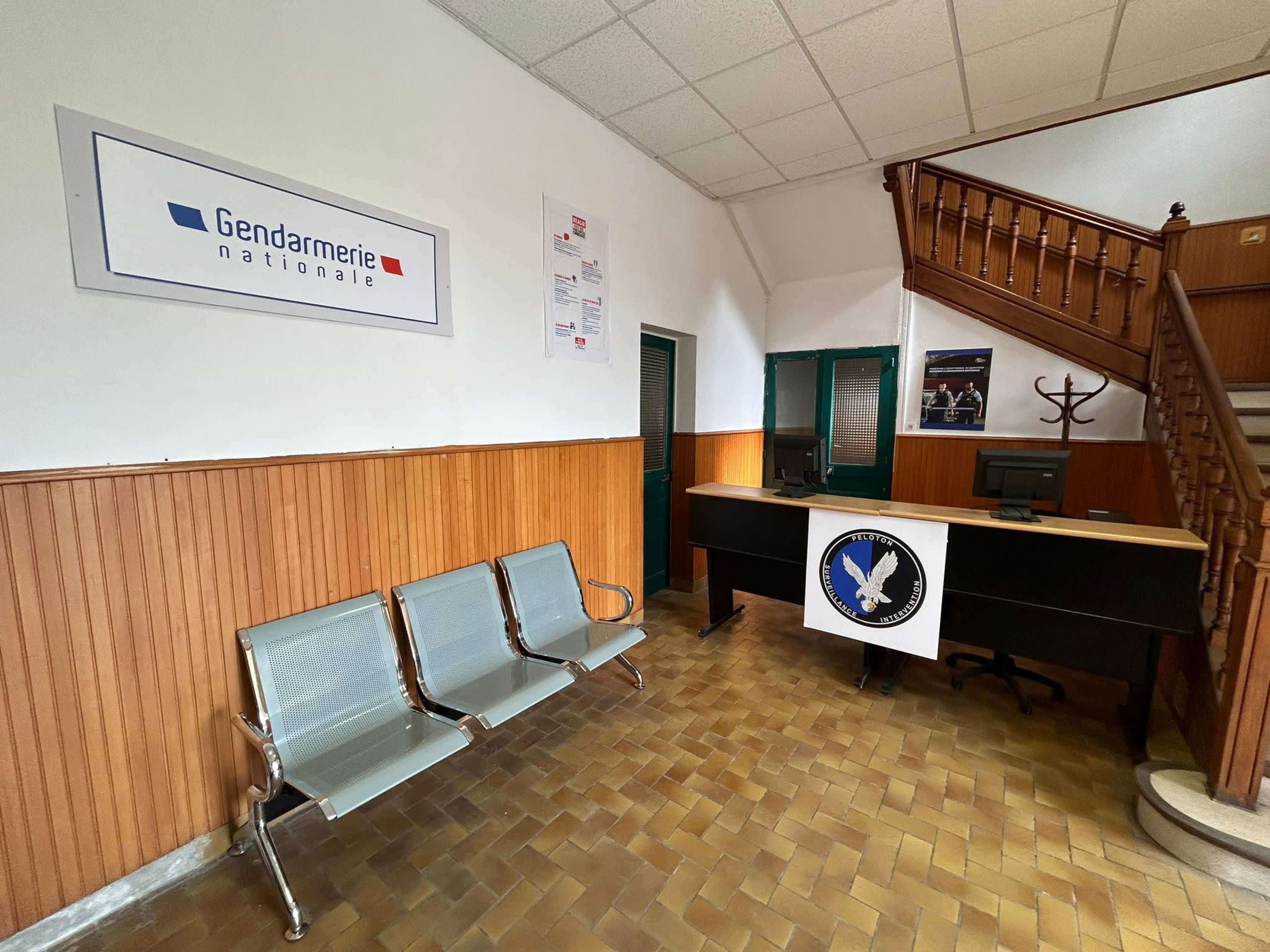 The interior of a gendarmerie station with metal benches, a reception desk displaying a logo, and wooden paneling on the walls.