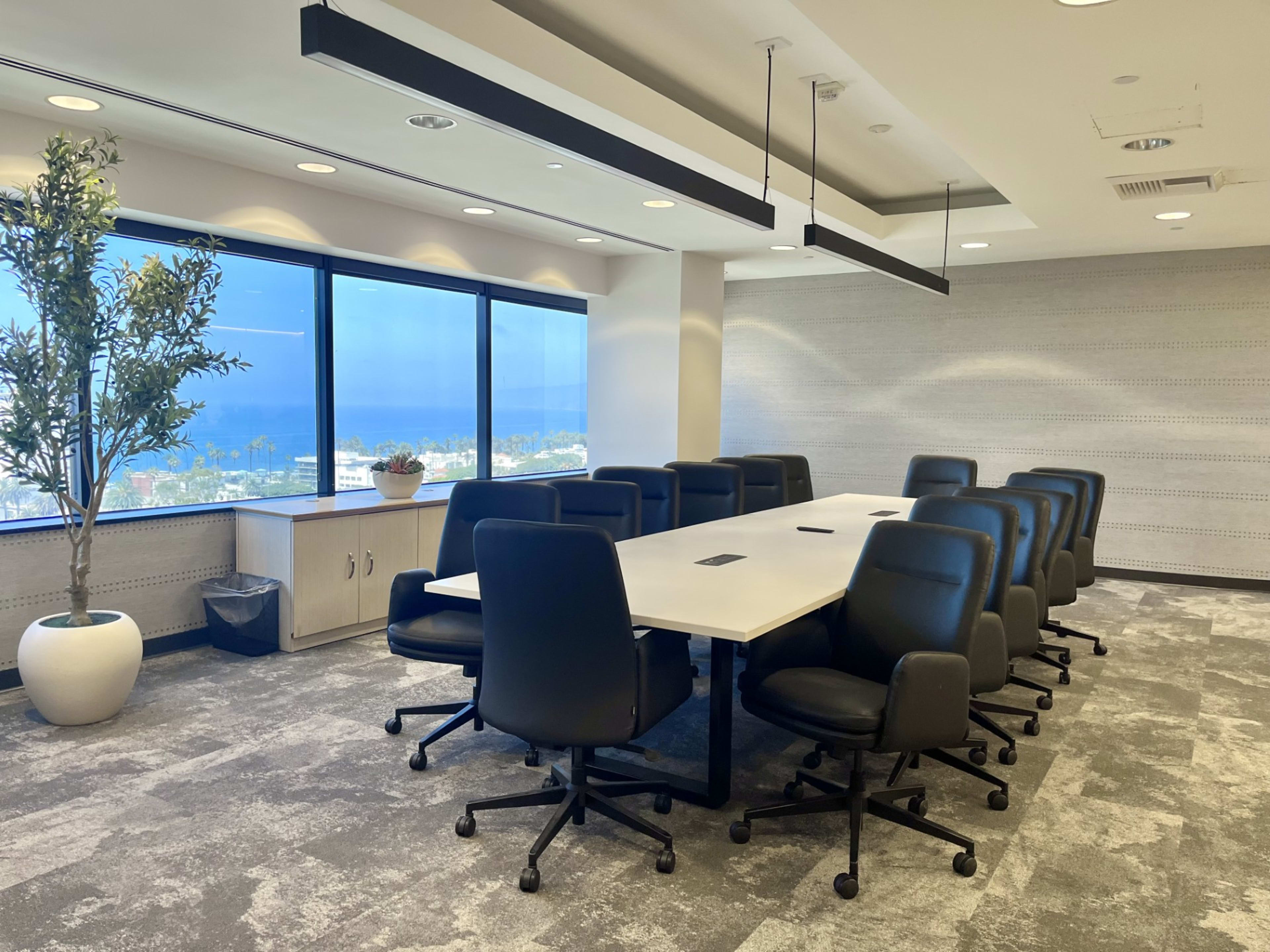 A modern conference room features a long white table surrounded by black chairs, with large windows offering a view of the ocean.