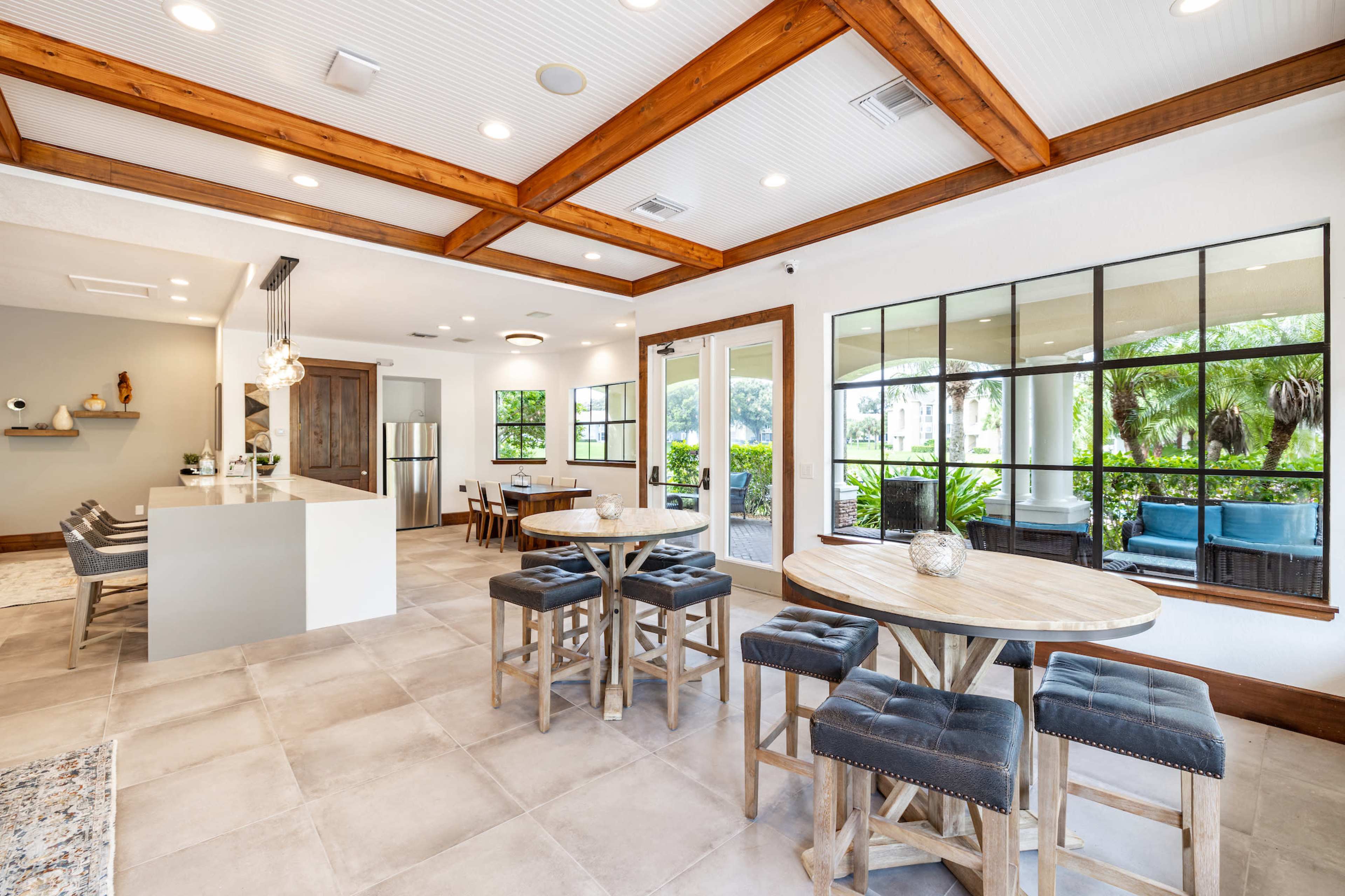 A modern dining area with wooden beams, circular tables, and a view of a shaded outdoor space through large windows.