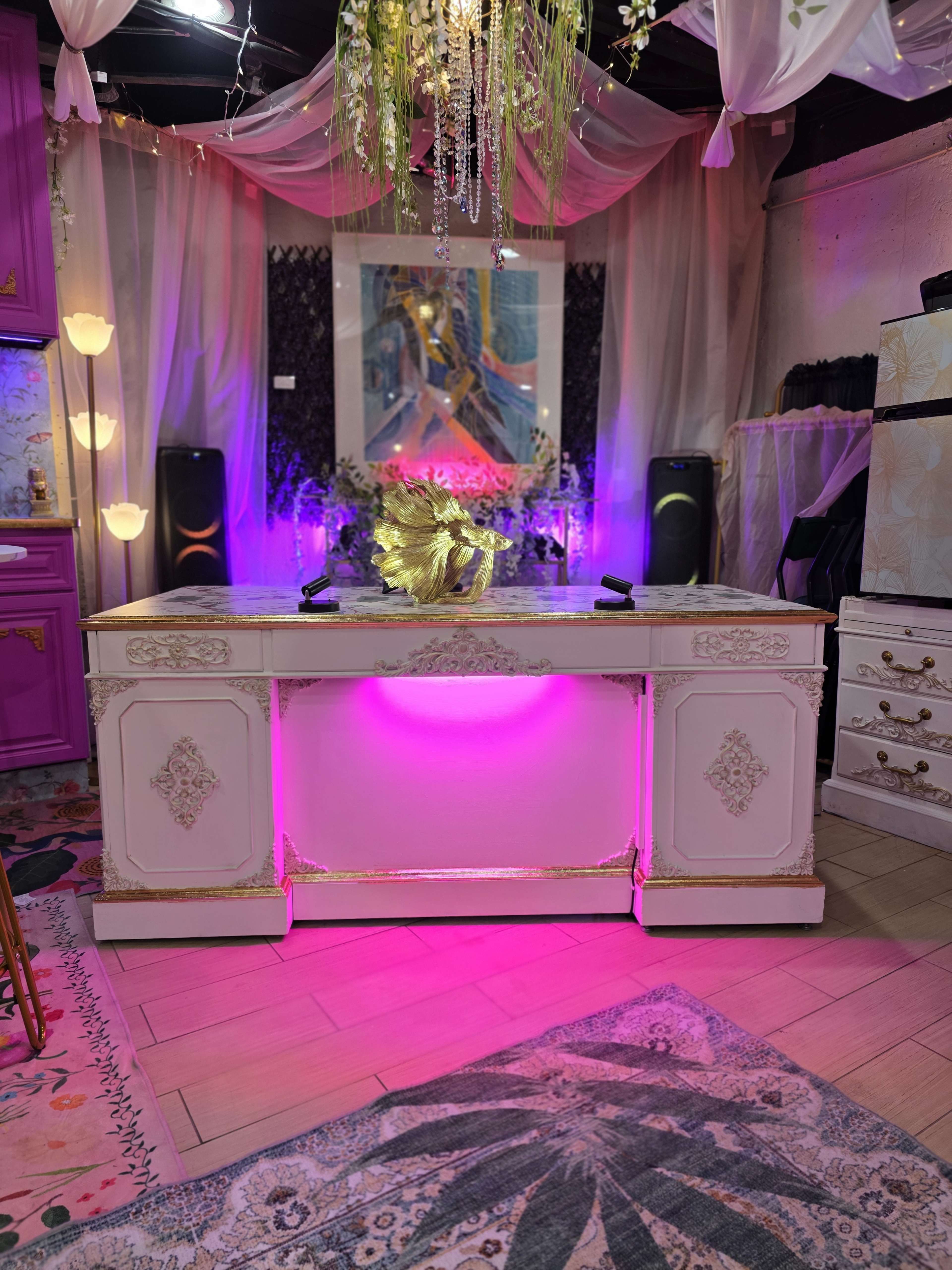 The image shows a decorated room featuring a white desk with pink lighting, surrounded by plants and colorful artwork.