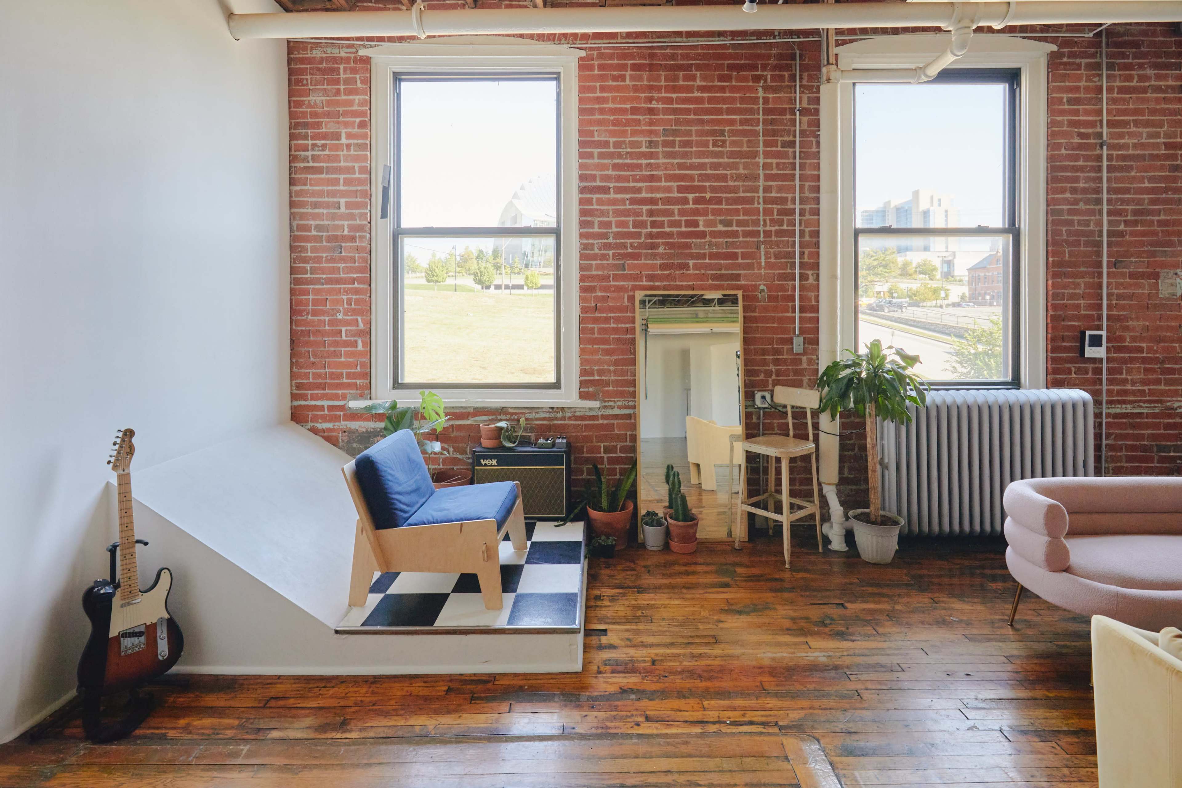 A spacious room featuring exposed brick walls, large windows, wooden floors, and a combination of minimalist furniture including a blue sofa, a wooden chair, and a small table, with greenery and a radiator adding to the decor.