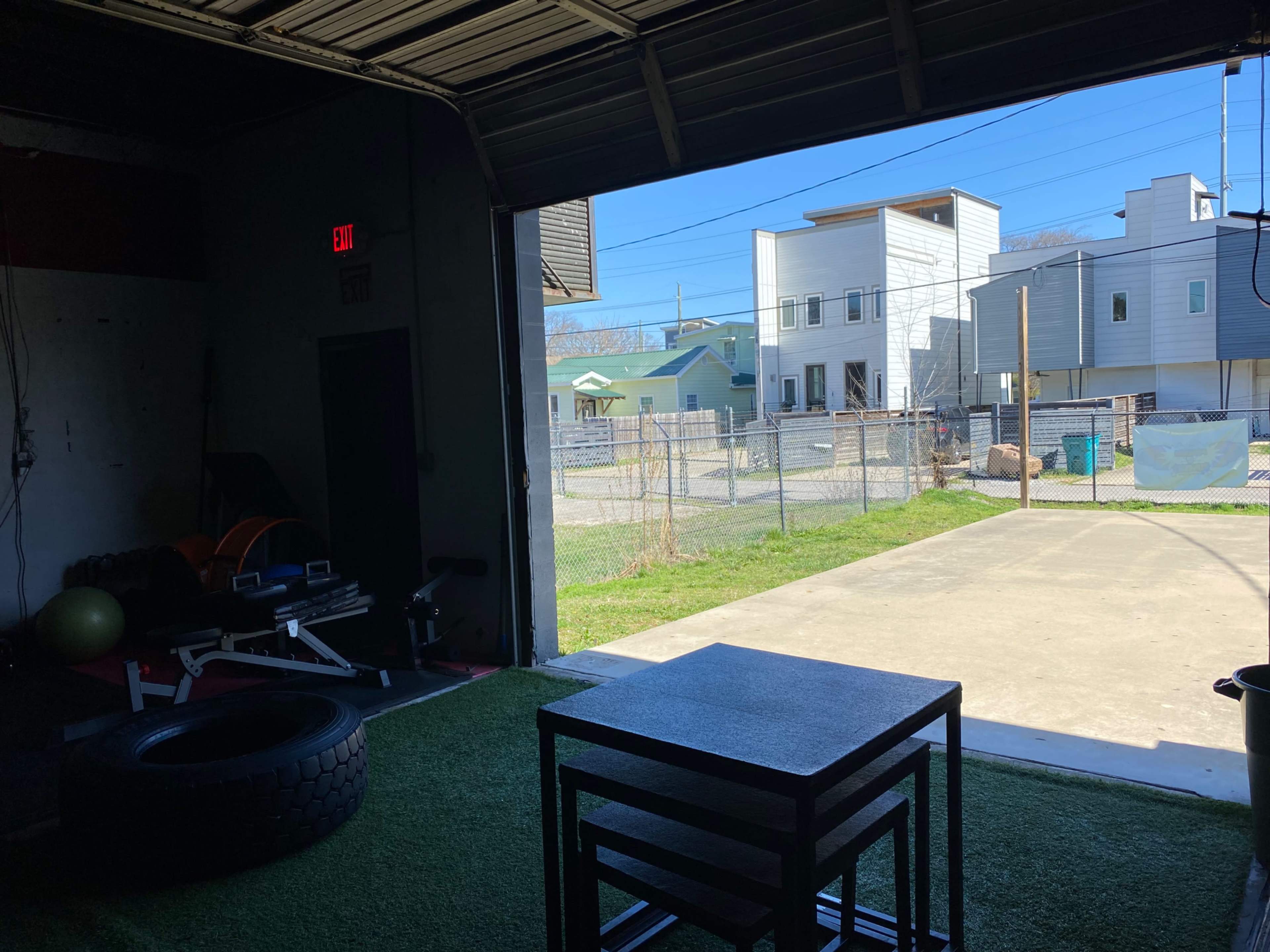 The interior of a gym with exercise equipment and a view of a fenced outdoor area and nearby buildings through an open garage door.