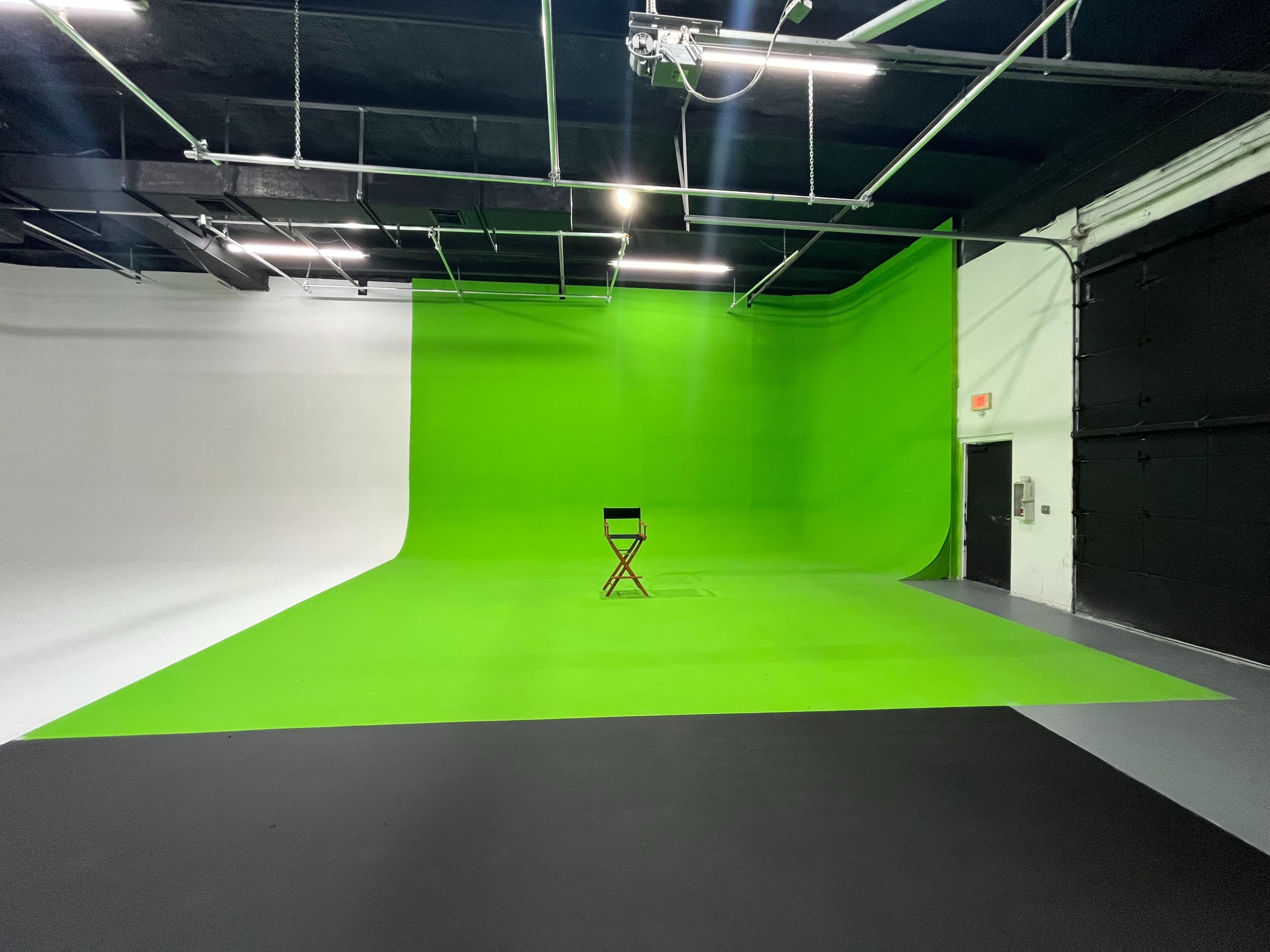 The image shows a photography studio with a green screen backdrop, a white wall, and a wooden director's chair positioned on a gray floor.