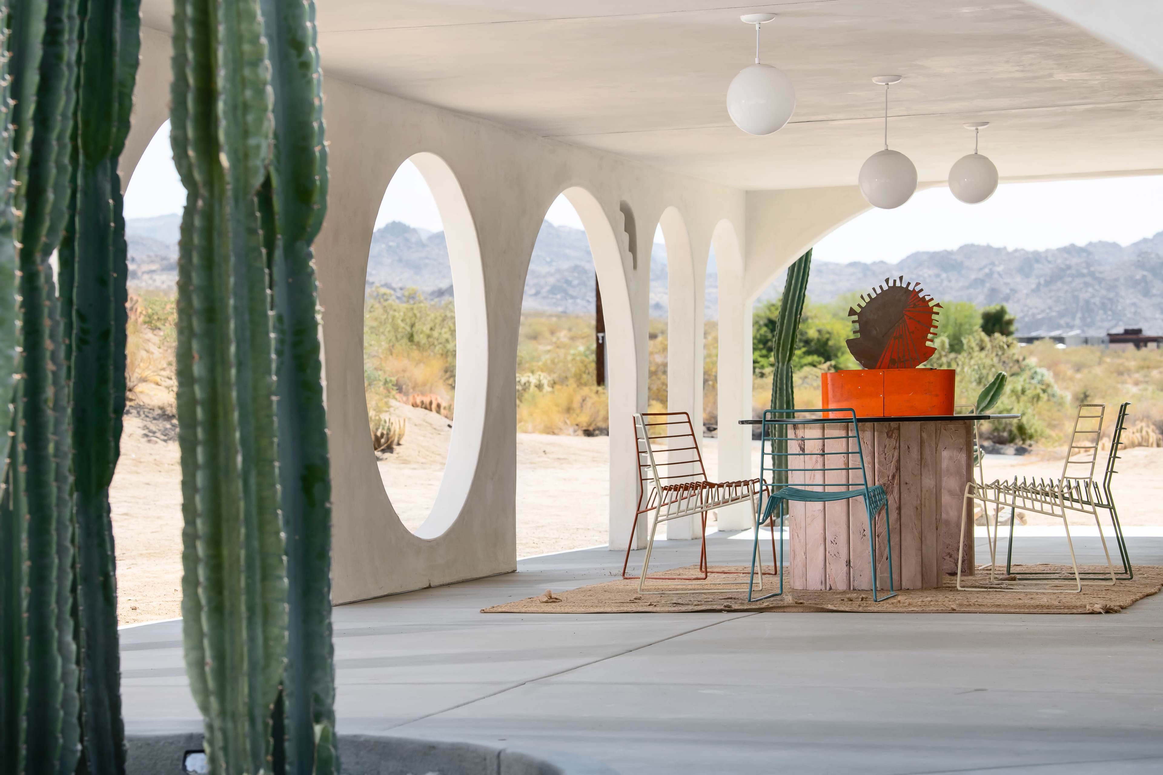 A modern outdoor seating area features a wooden table surrounded by wire chairs, framed by large arches and cacti in the foreground.