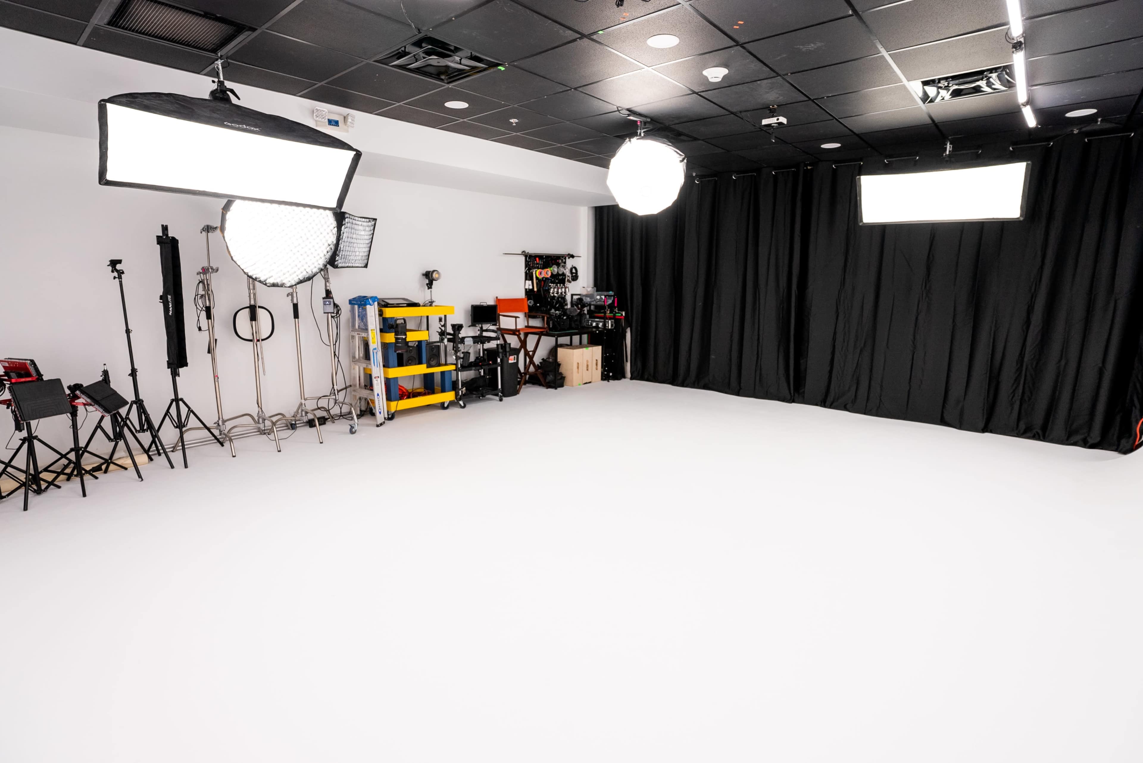 The image shows a photo studio with white flooring, various lighting equipment, and a black curtain backdrop.