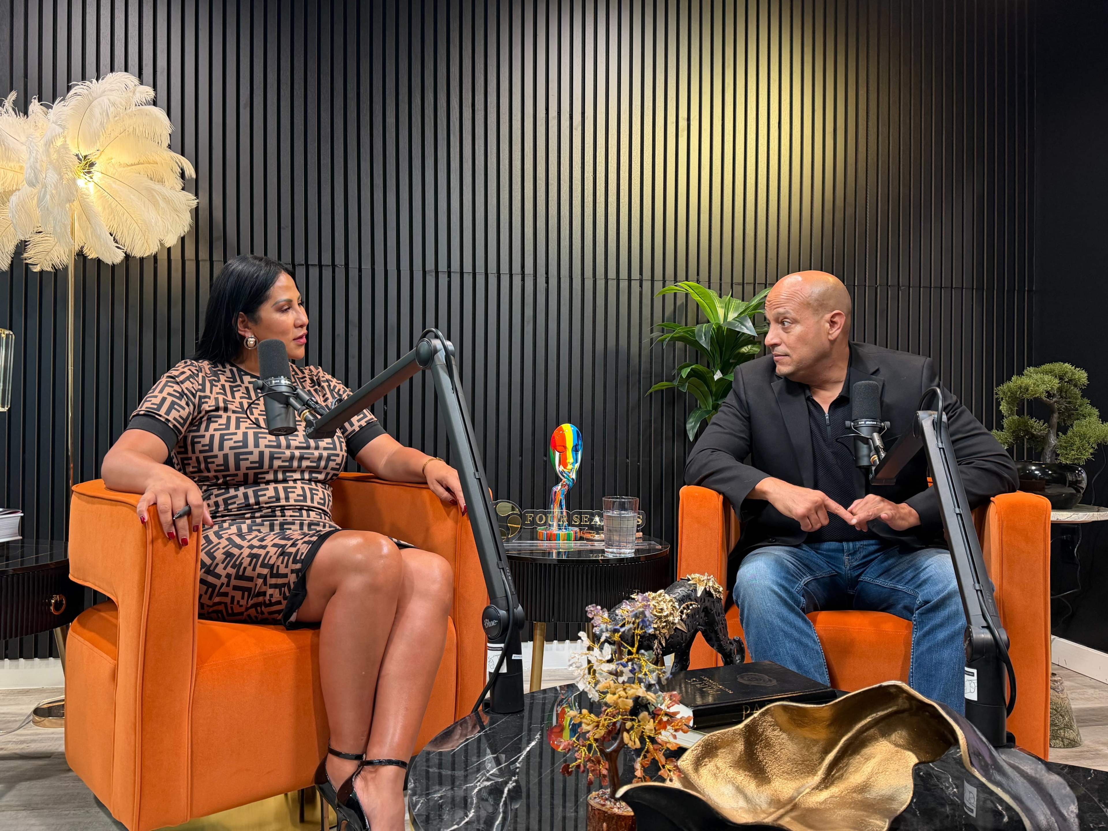 Two people are seated in orange armchairs during a discussion in a modern studio with dark walls and decorative elements.