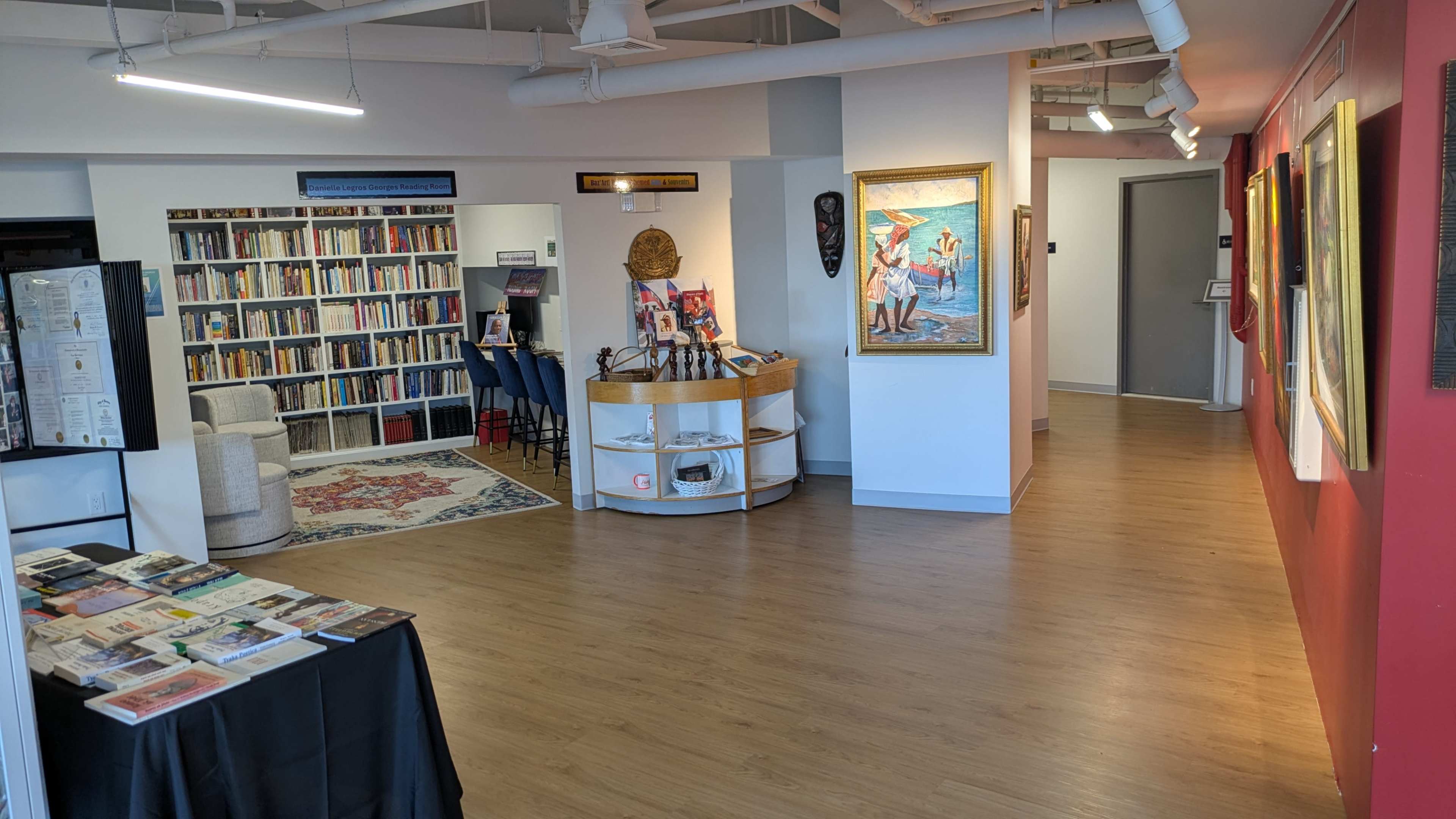 The image shows a spacious interior of an art gallery or community space, featuring shelves of books, a display table with brochures, and artwork hanging on the walls.