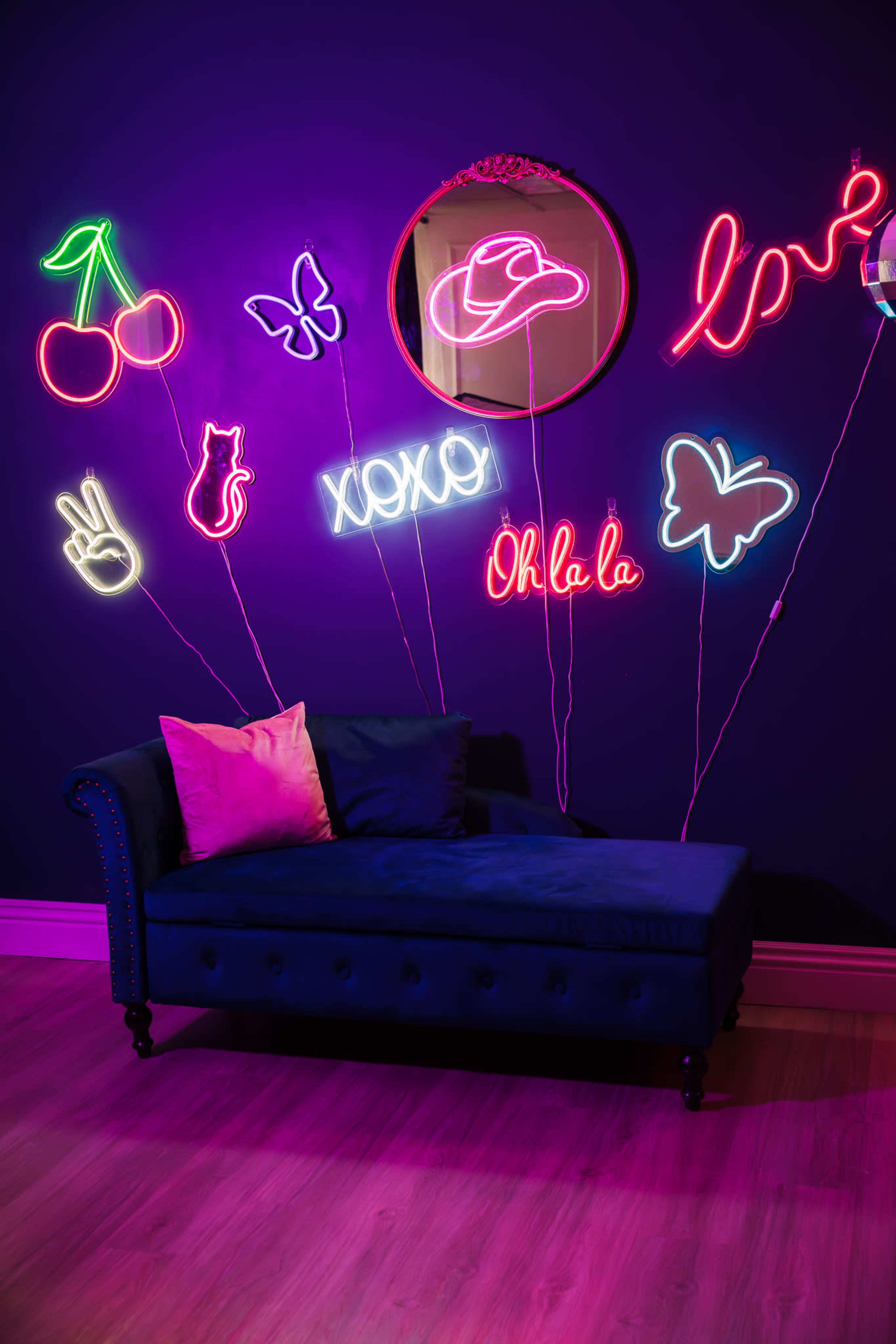 A plush dark blue couch is against a purple wall adorned with various neon signs, including cherries, butterflies, and the word "Ohlala."