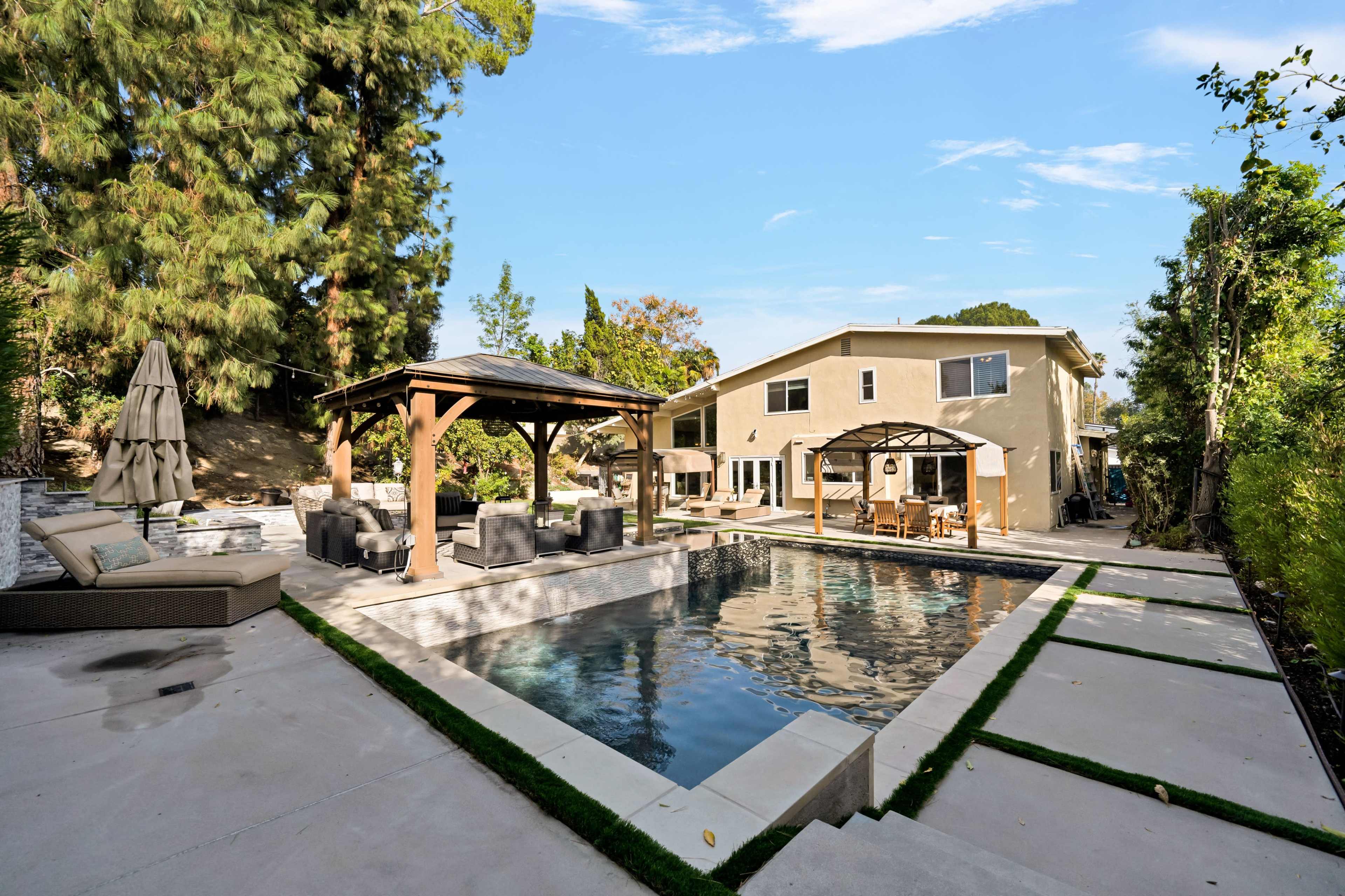 A landscaped backyard features a swimming pool surrounded by seating areas, a gazebo, and a two-story house.