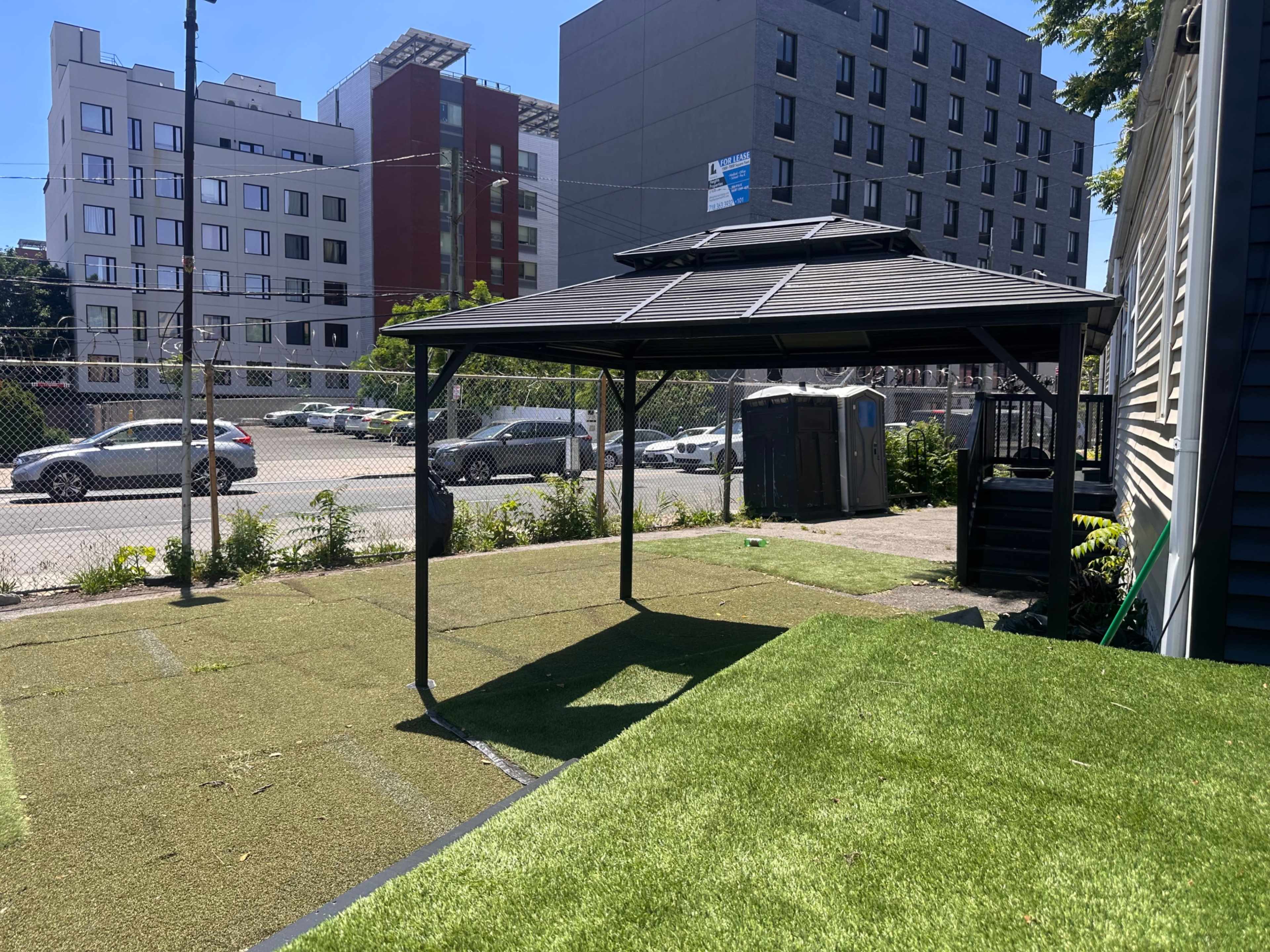 The image shows a shaded gazebo in a grassy area with buildings in the background.