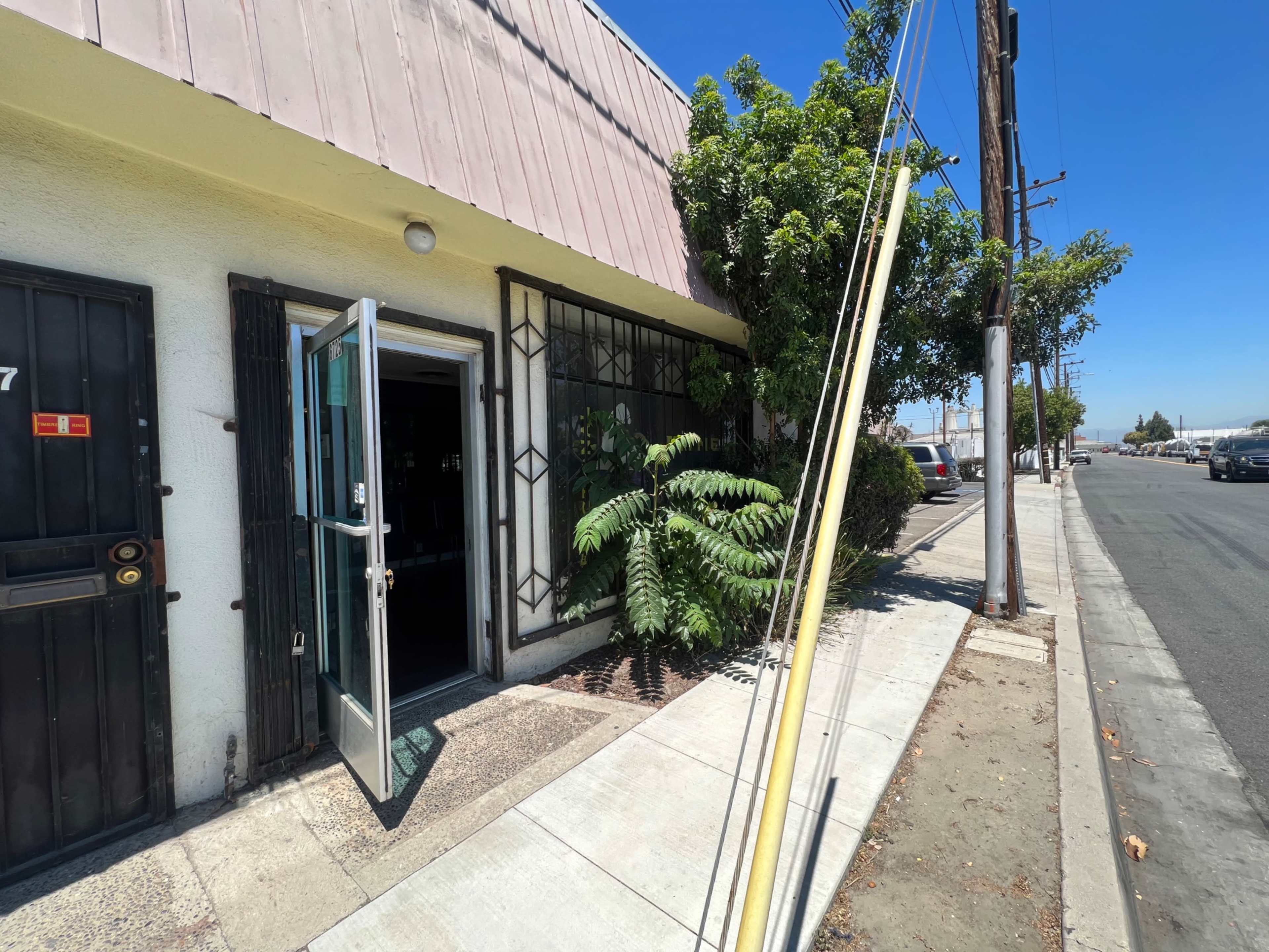 The image shows the entrance of a building with an open door, surrounded by greenery and positioned next to a sidewalk and street.