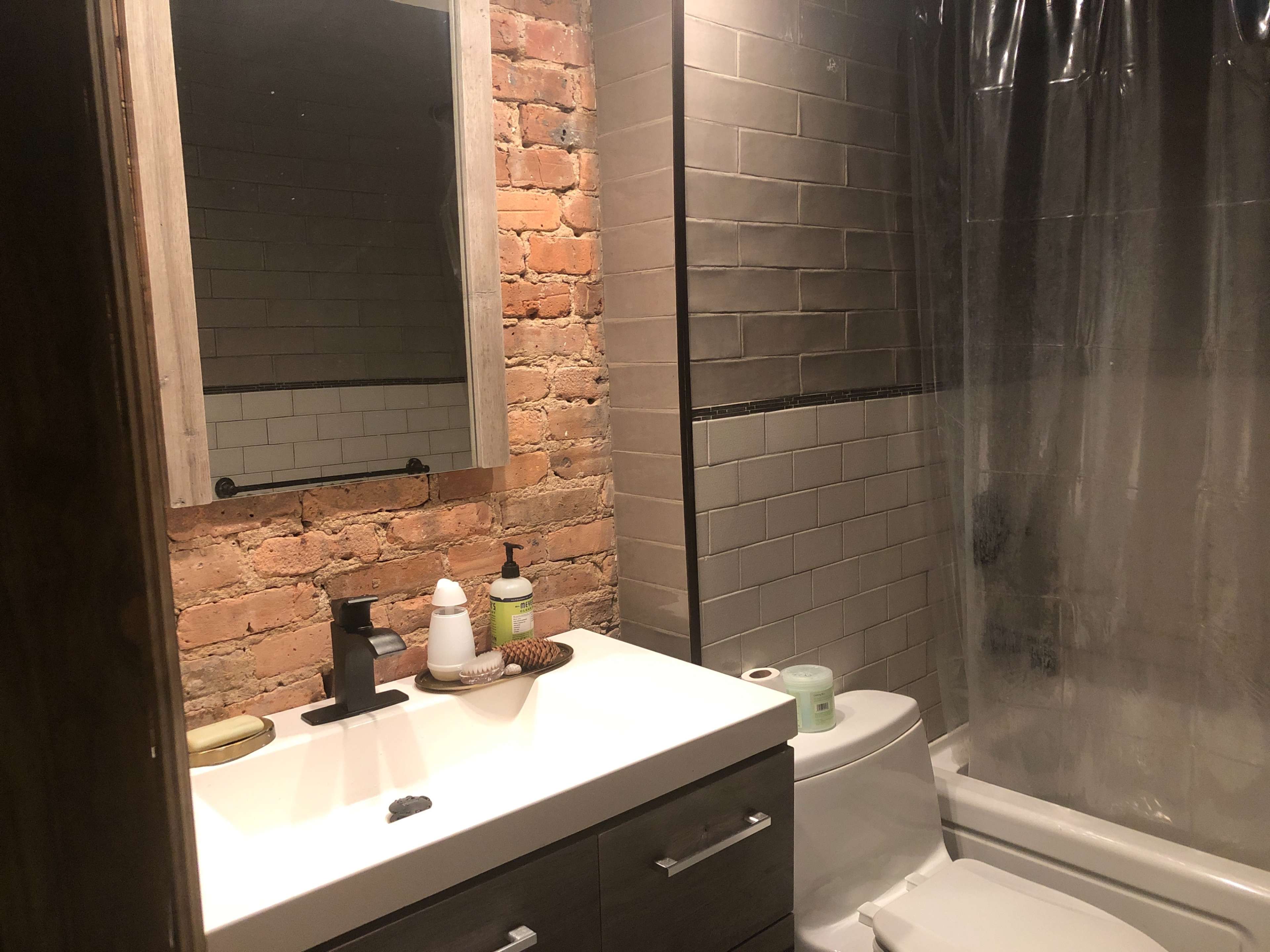 A bathroom featuring exposed brick walls, a modern sink with a mirror above it, a shower with a clear curtain, and a toilet placed next to the tub.