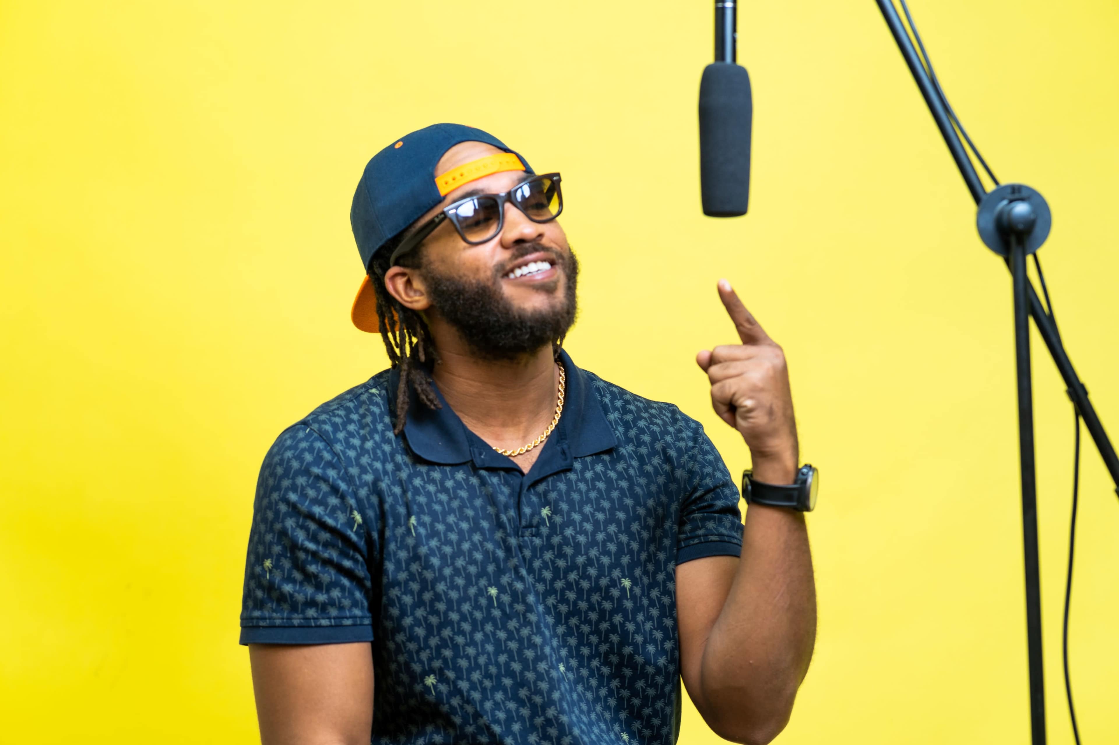 A man with long hair in braids wearing sunglasses and a cap points upward in front of a microphone against a yellow background.