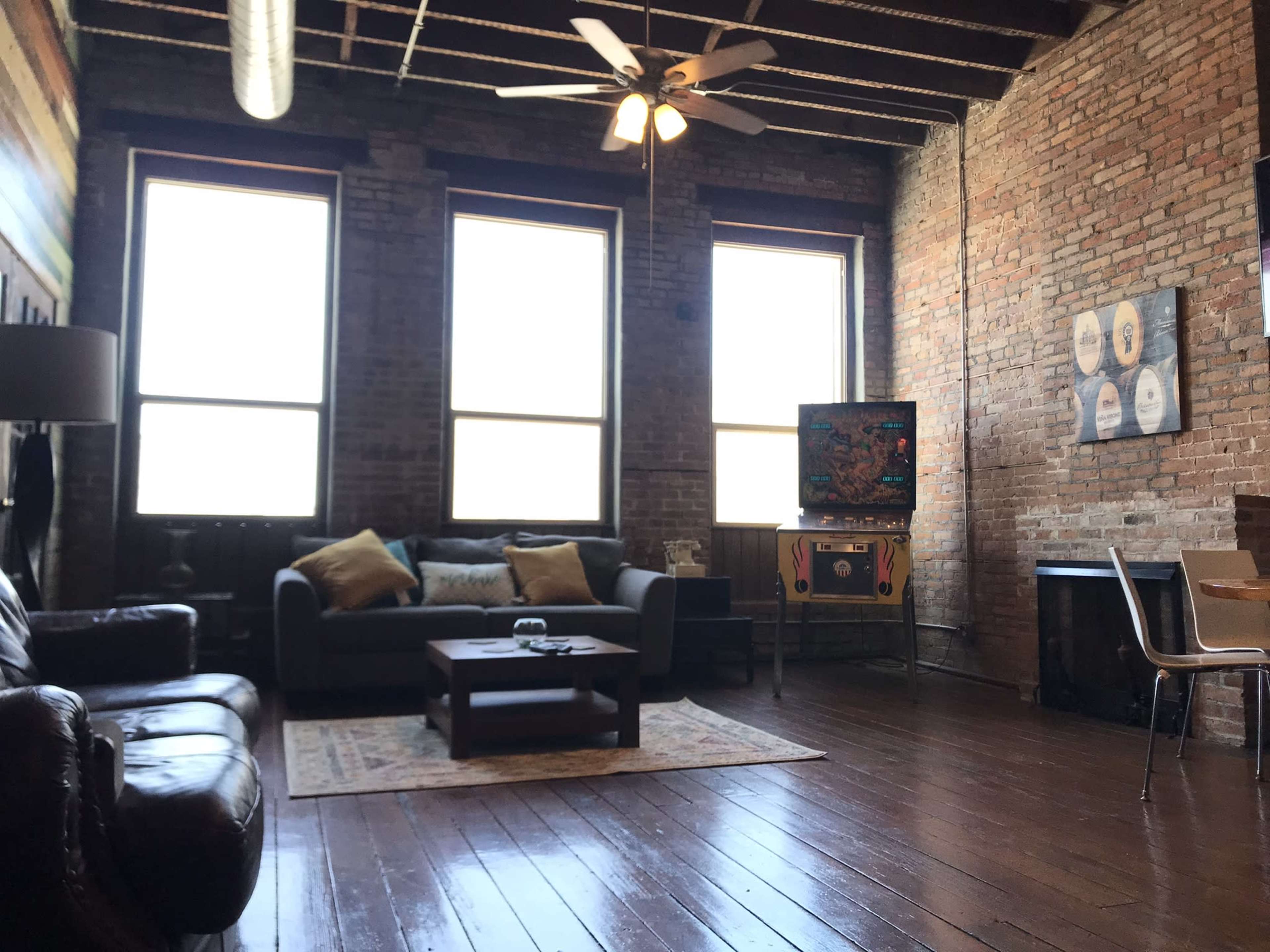 The image shows a spacious, industrial-style living room with large windows, exposed brick walls, a leather sofa, and a wooden coffee table.