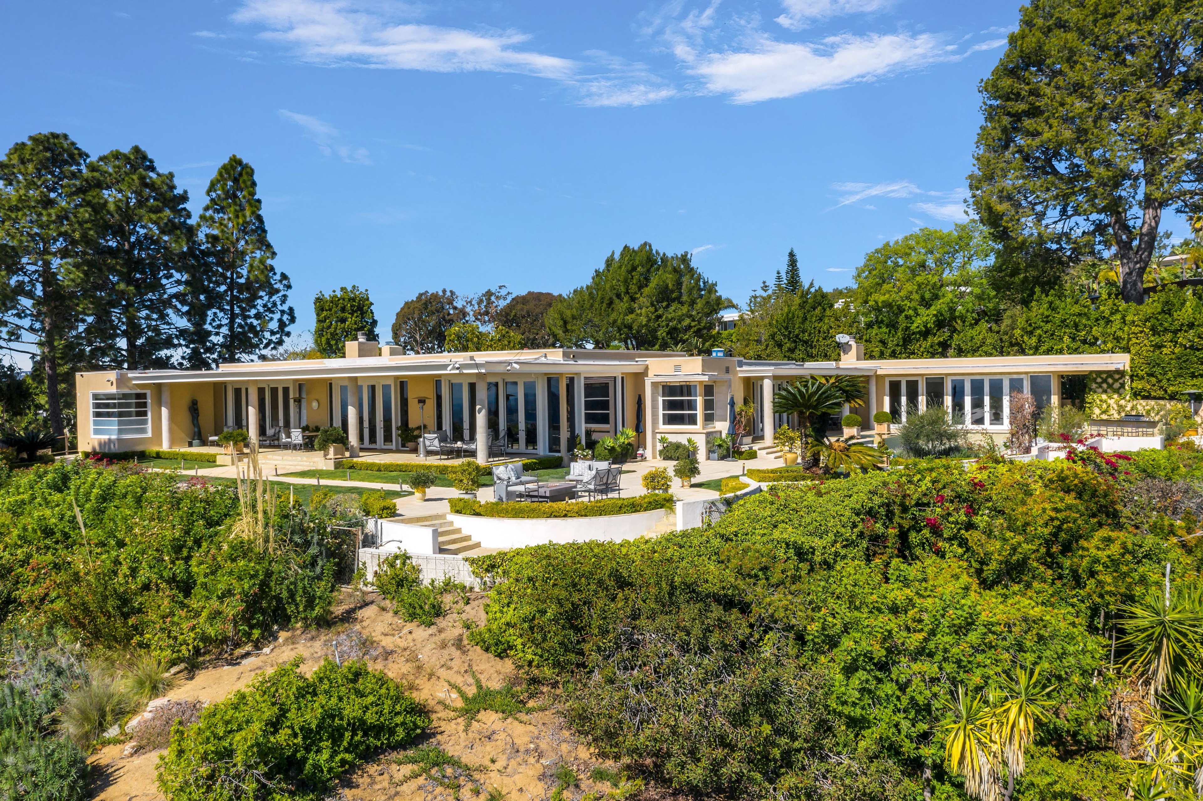 A single-story modern house with large windows is surrounded by lush landscaping and trees under a clear blue sky.