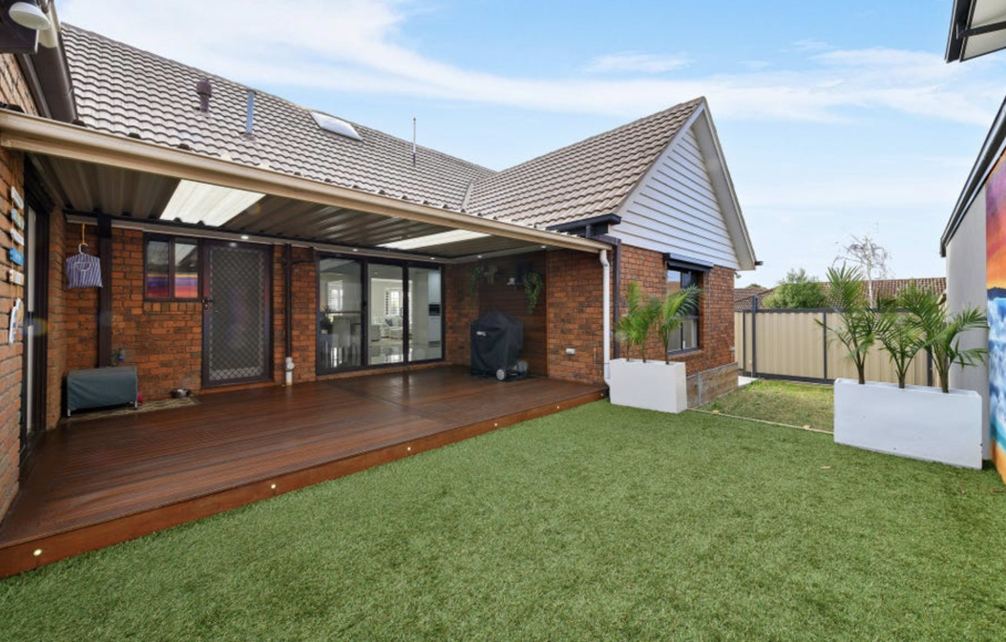 The image shows a backyard with a wooden deck, artificial grass, and a brick house featuring a covered patio area.
