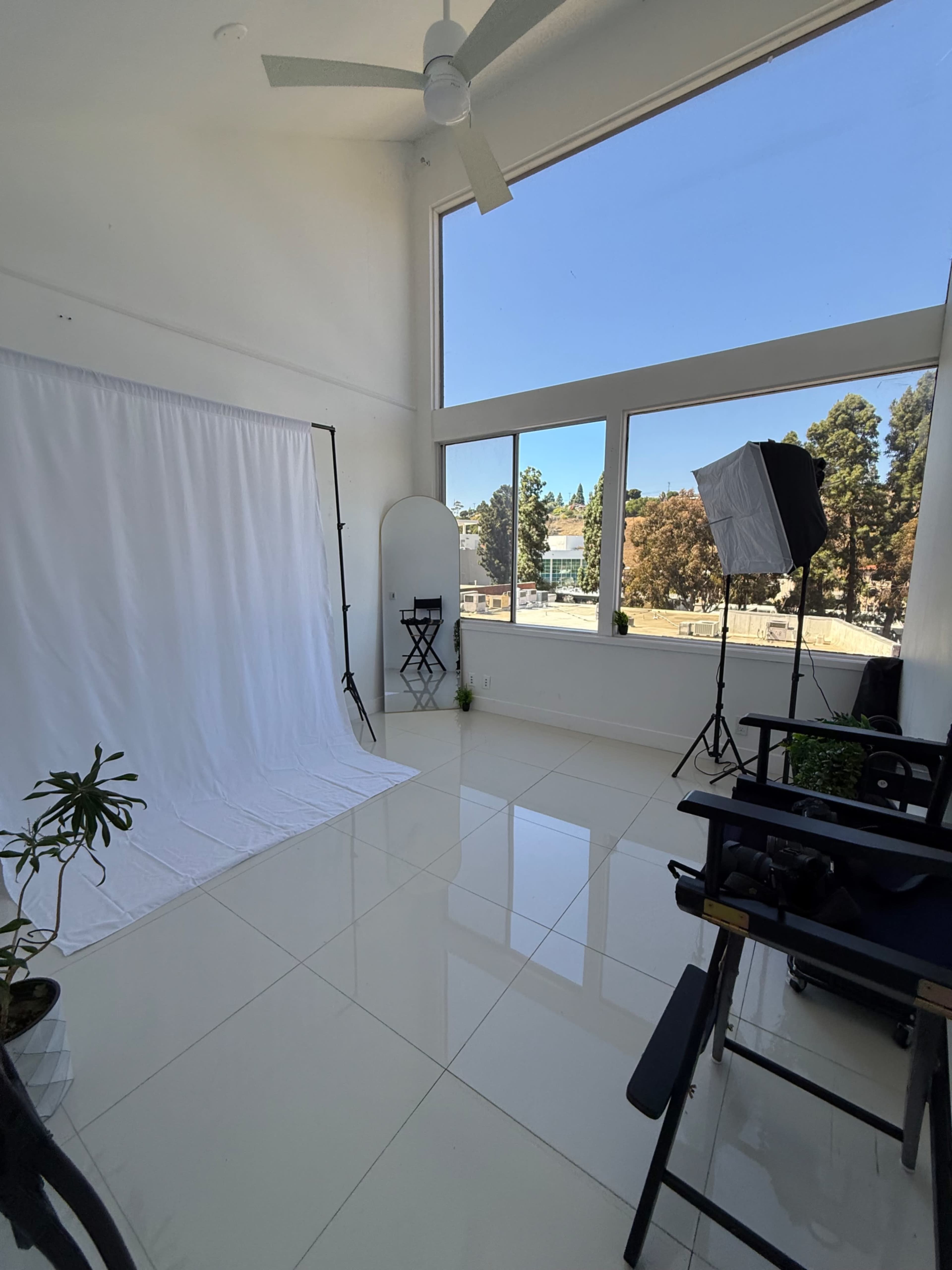 The image shows a bright, empty photography studio with a large window, white backdrop, studio lights, and seating arrangements.