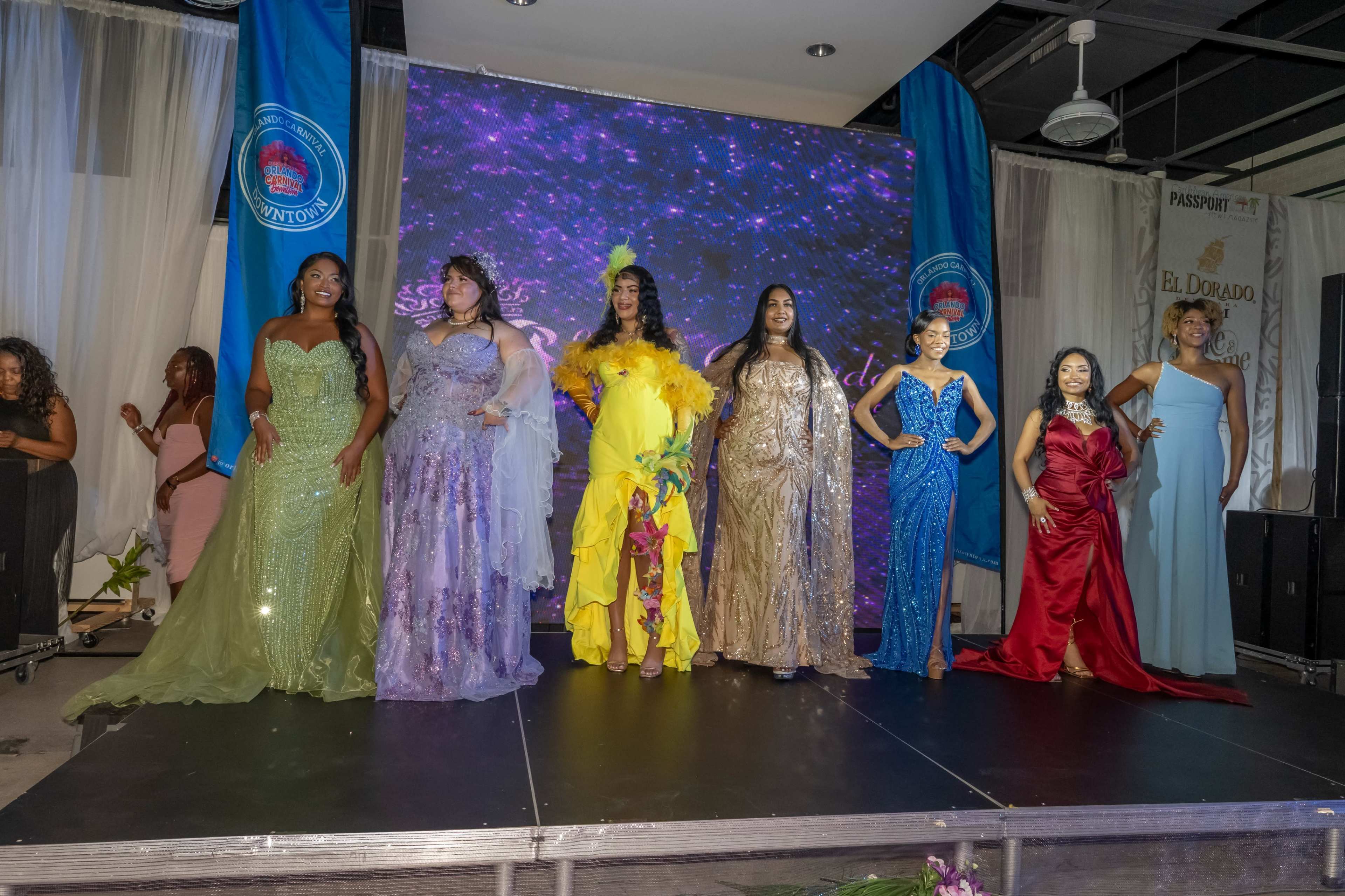 A group of seven women in various colorful evening gowns poses on a stage during a fashion show event.