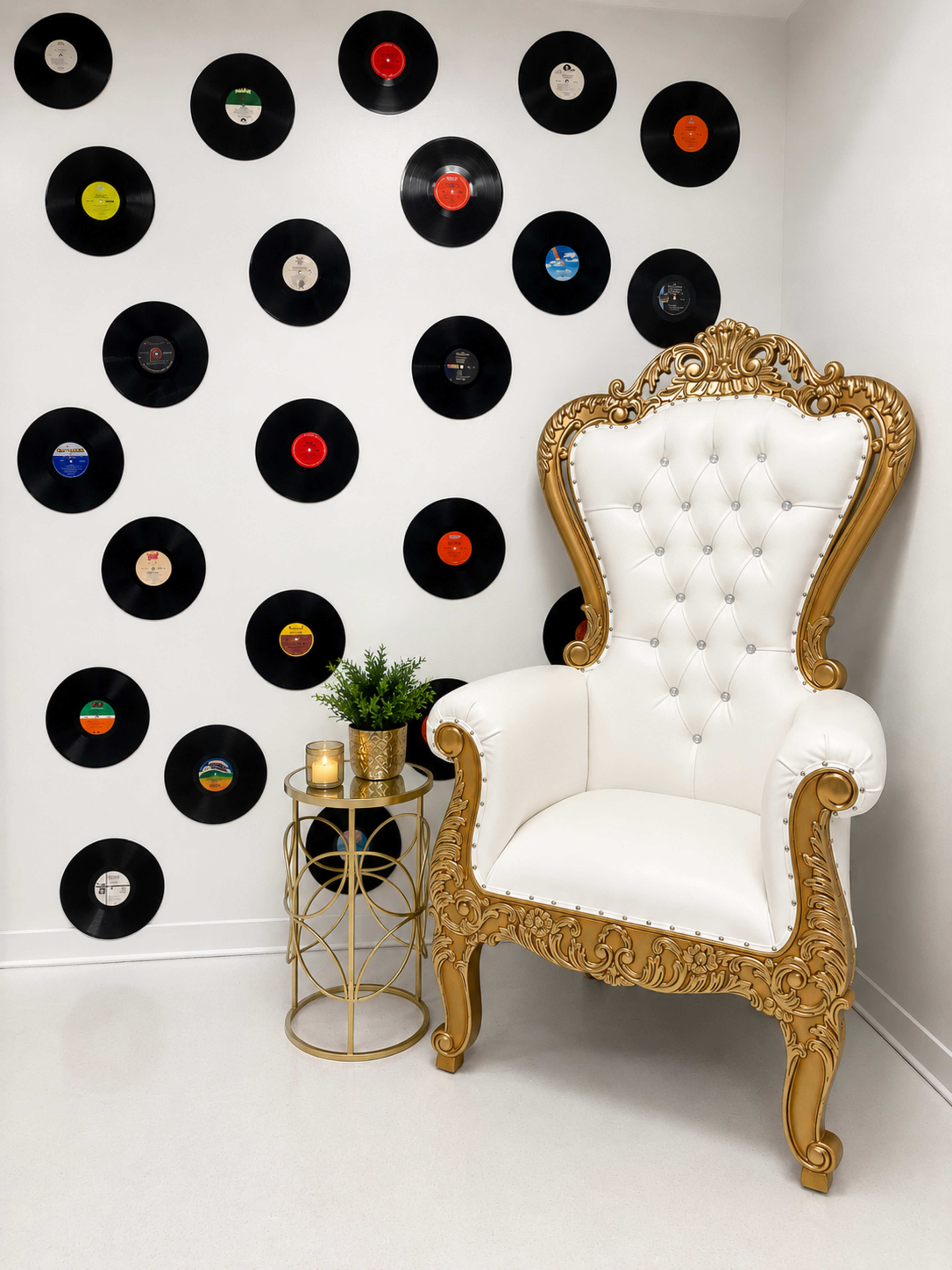 A vintage-style ornate white and gold chair is positioned next to a small gold side table adorned with a candle, against a white wall decorated with various vinyl records.