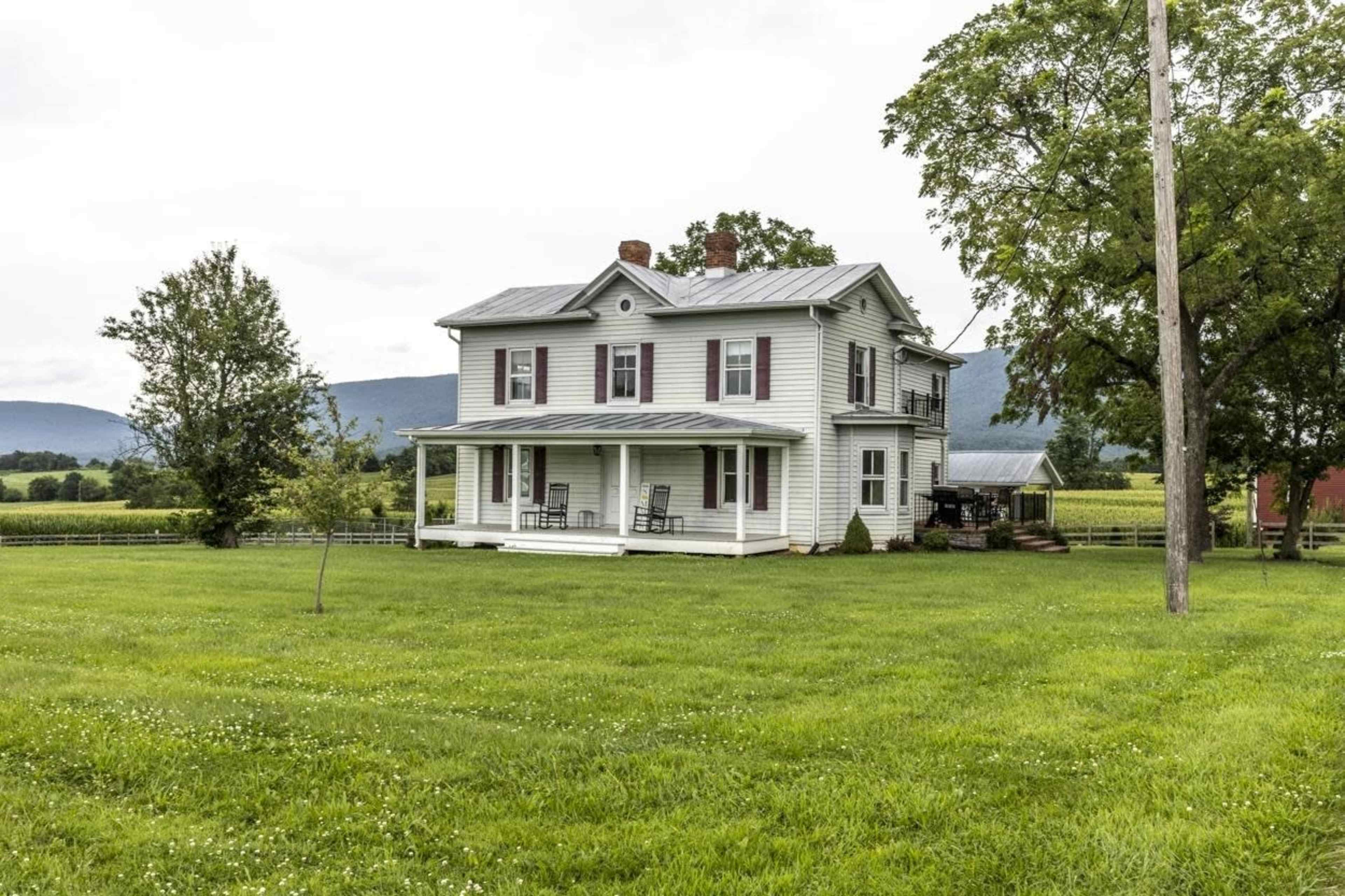 A two-story white farmhouse with a metal roof is situated on a grassy lawn surrounded by fields and trees.