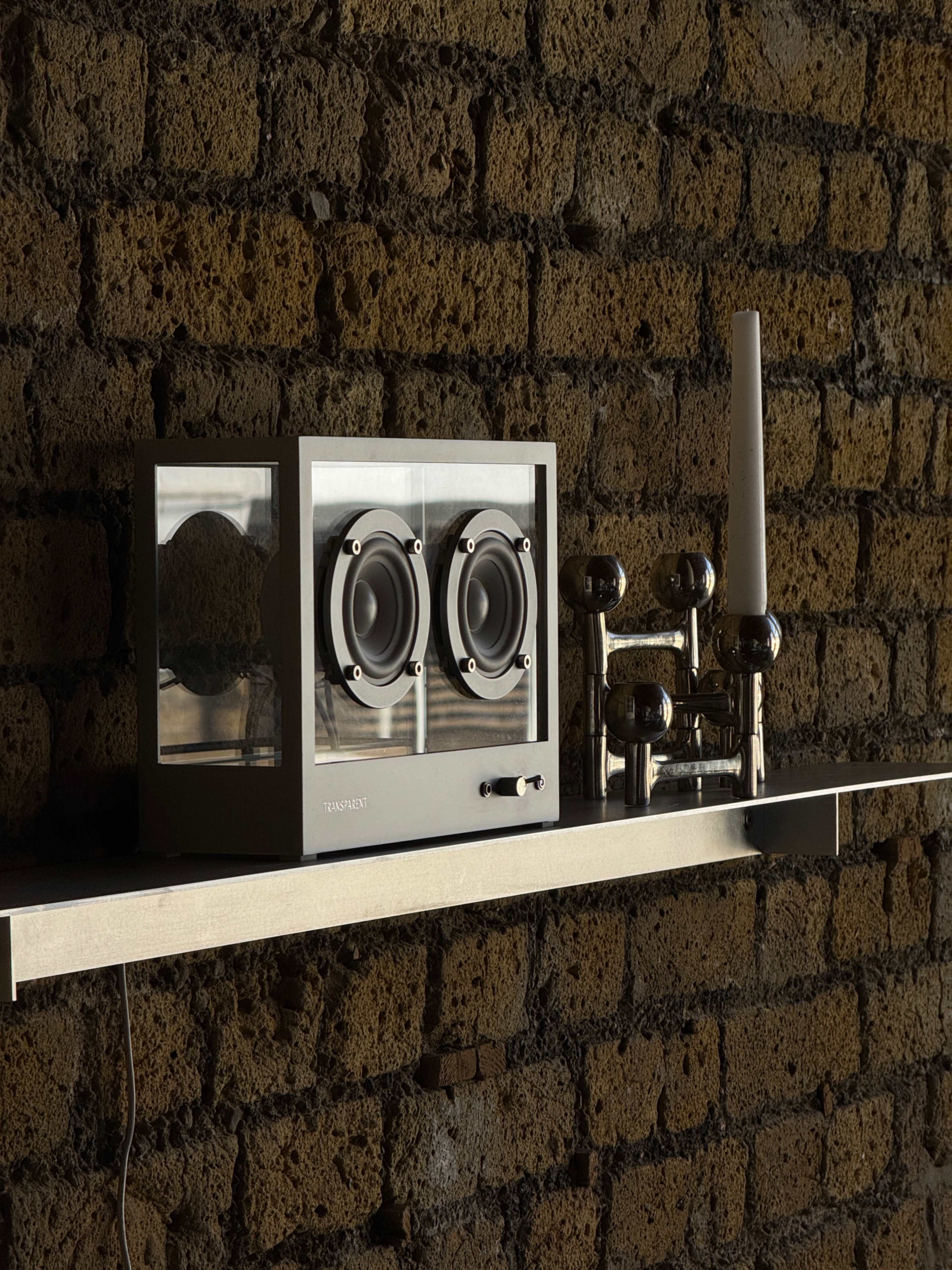 A modern speaker system with dual speakers and a minimalist design is displayed on a metal shelf against a brick wall.