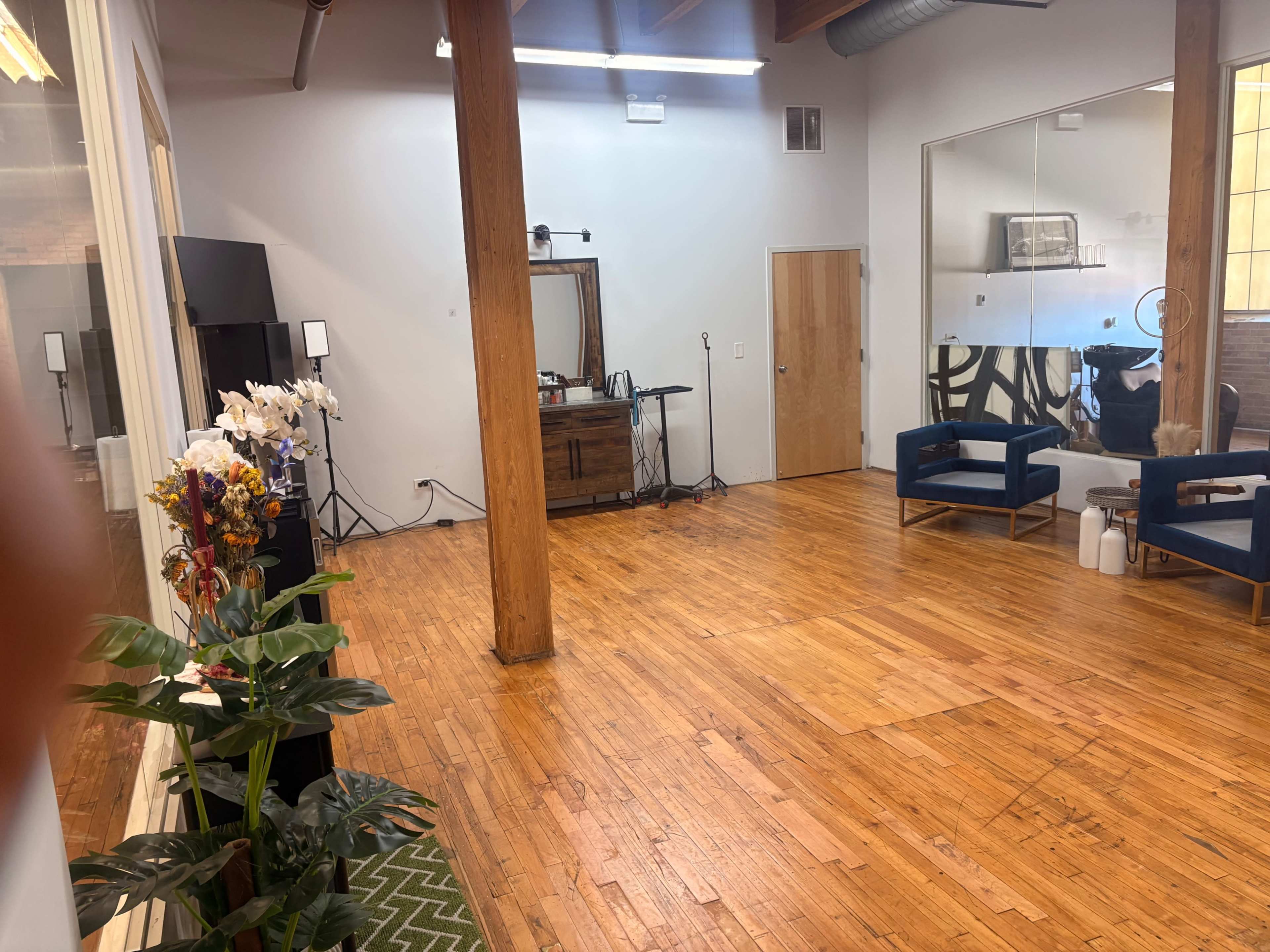 The image shows a spacious interior with wooden flooring, featuring two blue chairs, a decorative plant arrangement, and a large mirror on the wall.