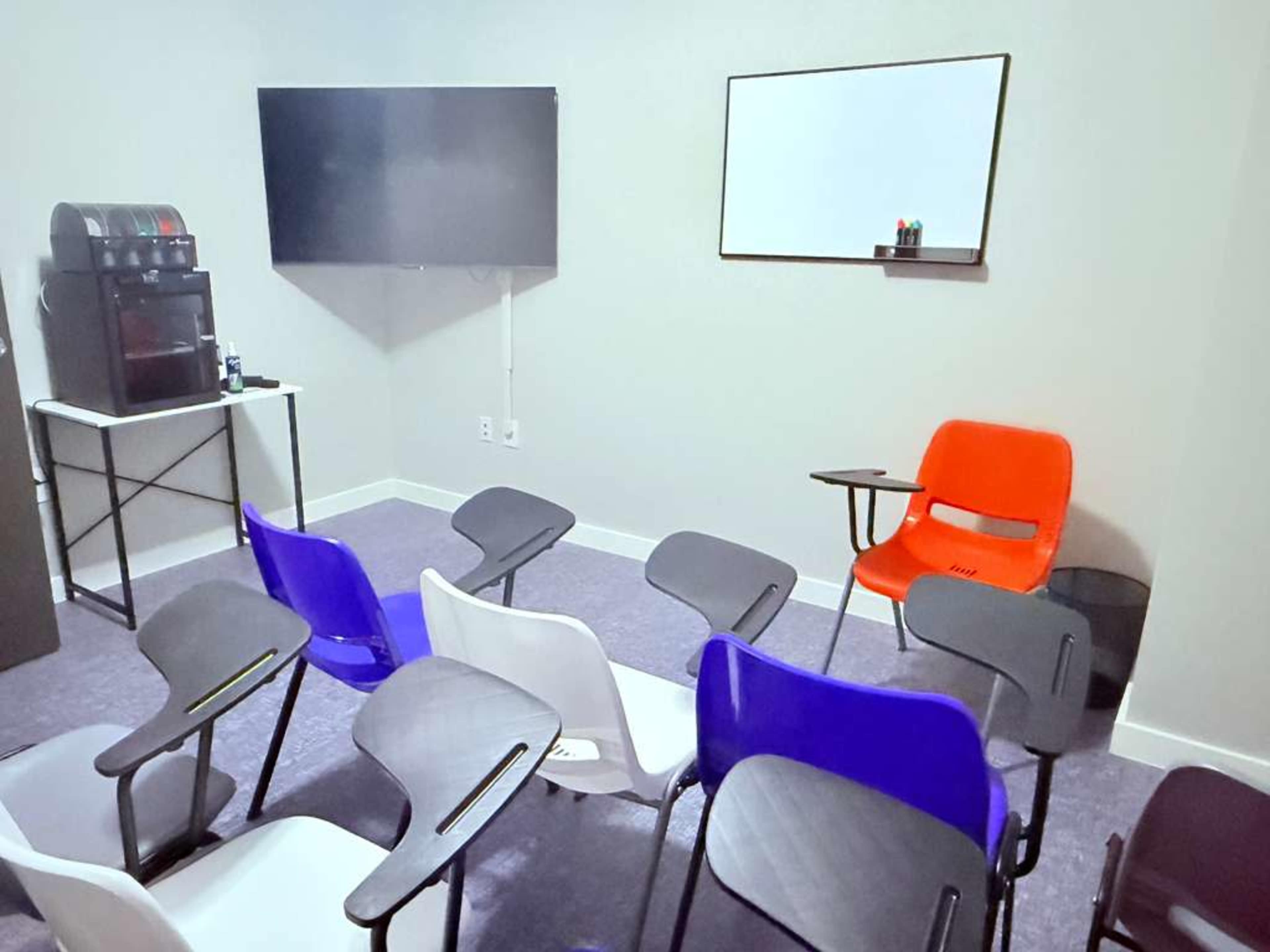 The image shows a small meeting room with several colorful chairs arranged around a tabletop, a whiteboard, and a television mounted on the wall.