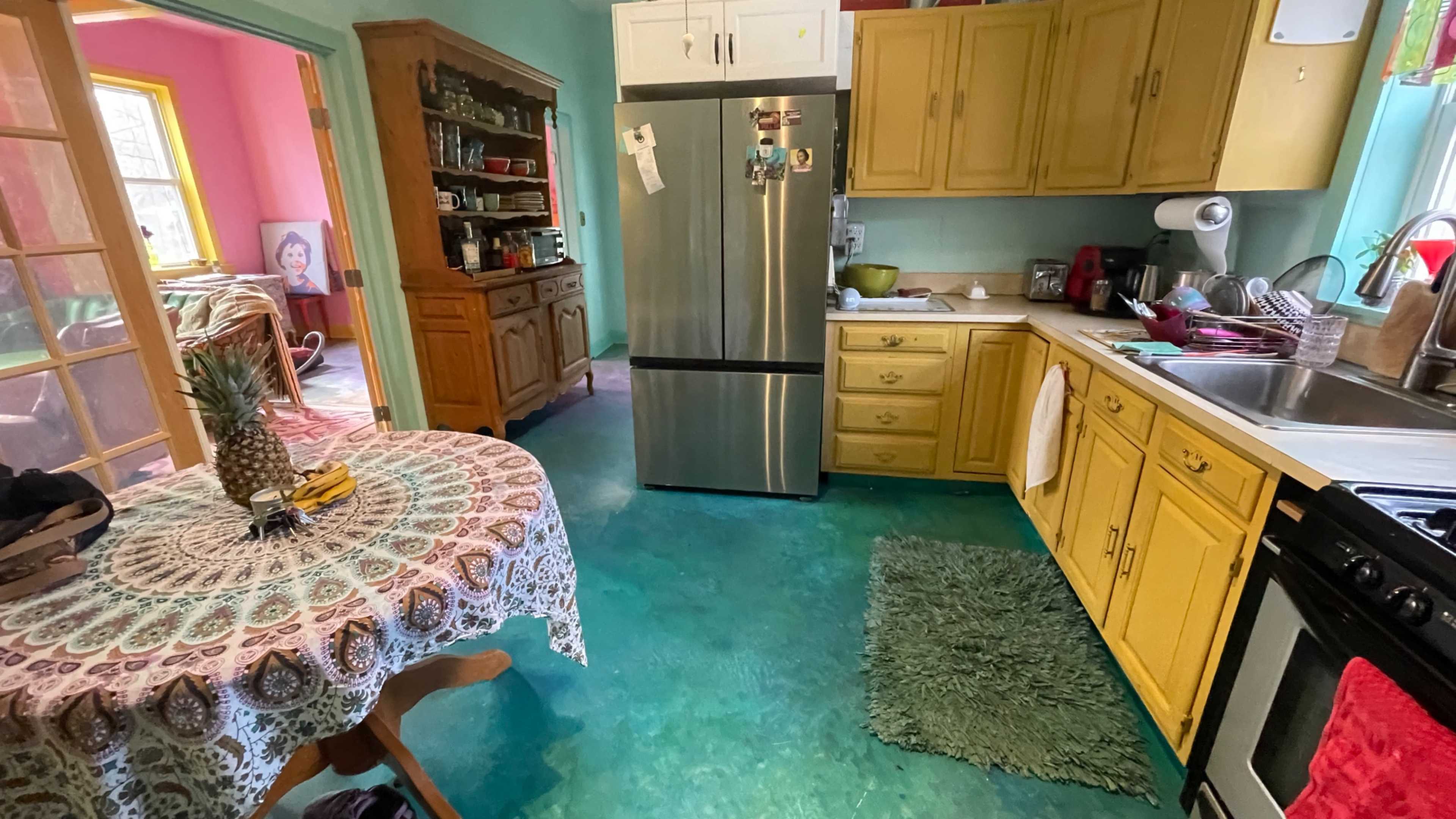 The image shows a colorful kitchen with yellow cabinets, a stainless steel refrigerator, and a circular table covered with a patterned tablecloth.