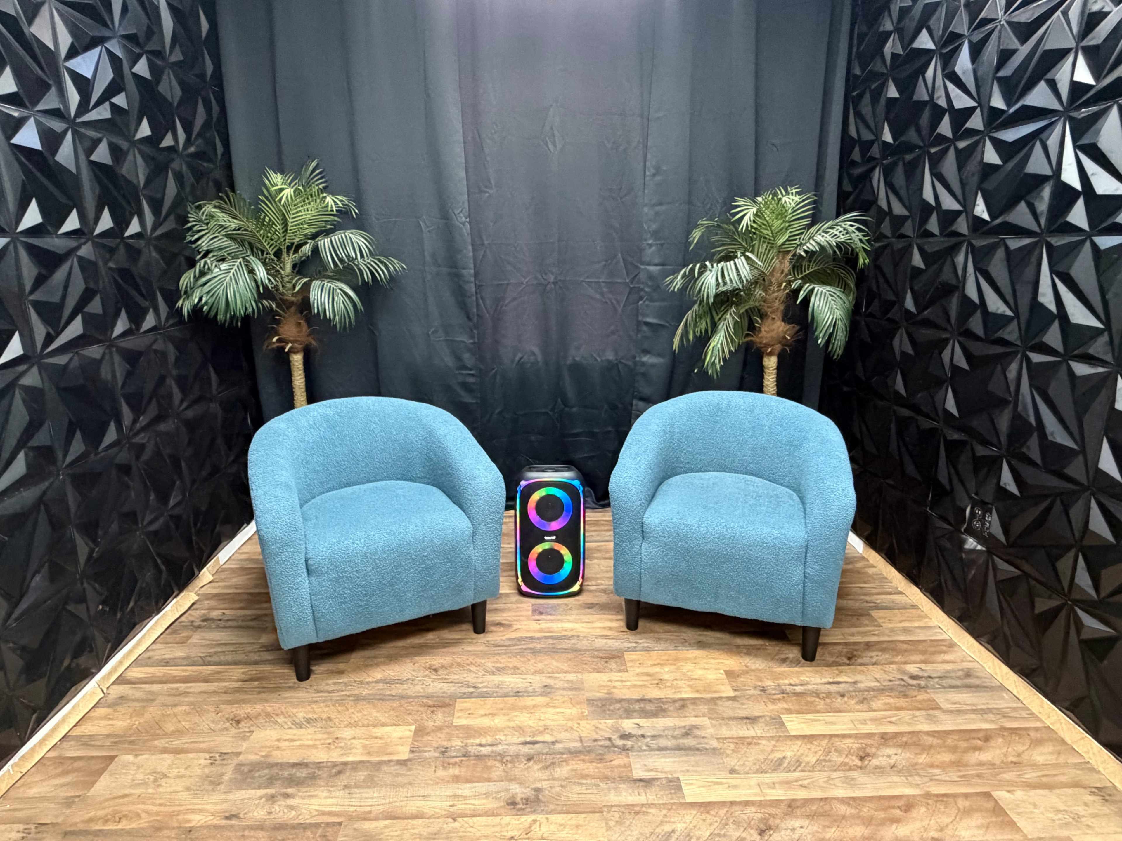 The image displays a small, modern seating area featuring two teal chairs with rounded backs flanking a brightly colored speaker, set against a backdrop of black geometric wall panels and two artificial palm trees.